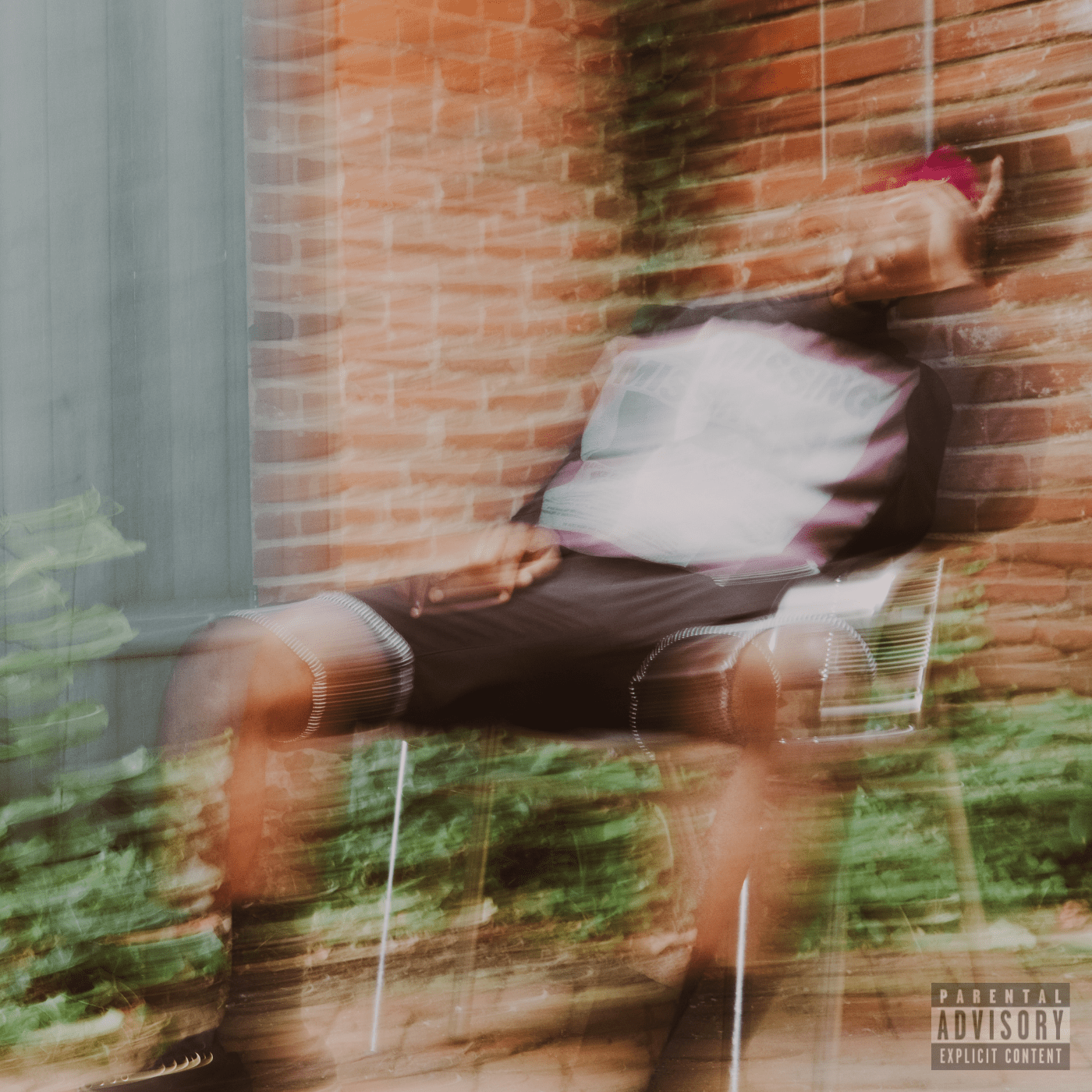 Blurry image of a person sitting on a chair, leaning against a brick wall with some greenery in the foreground, and a parental advisory sticker in the bottom right corner.