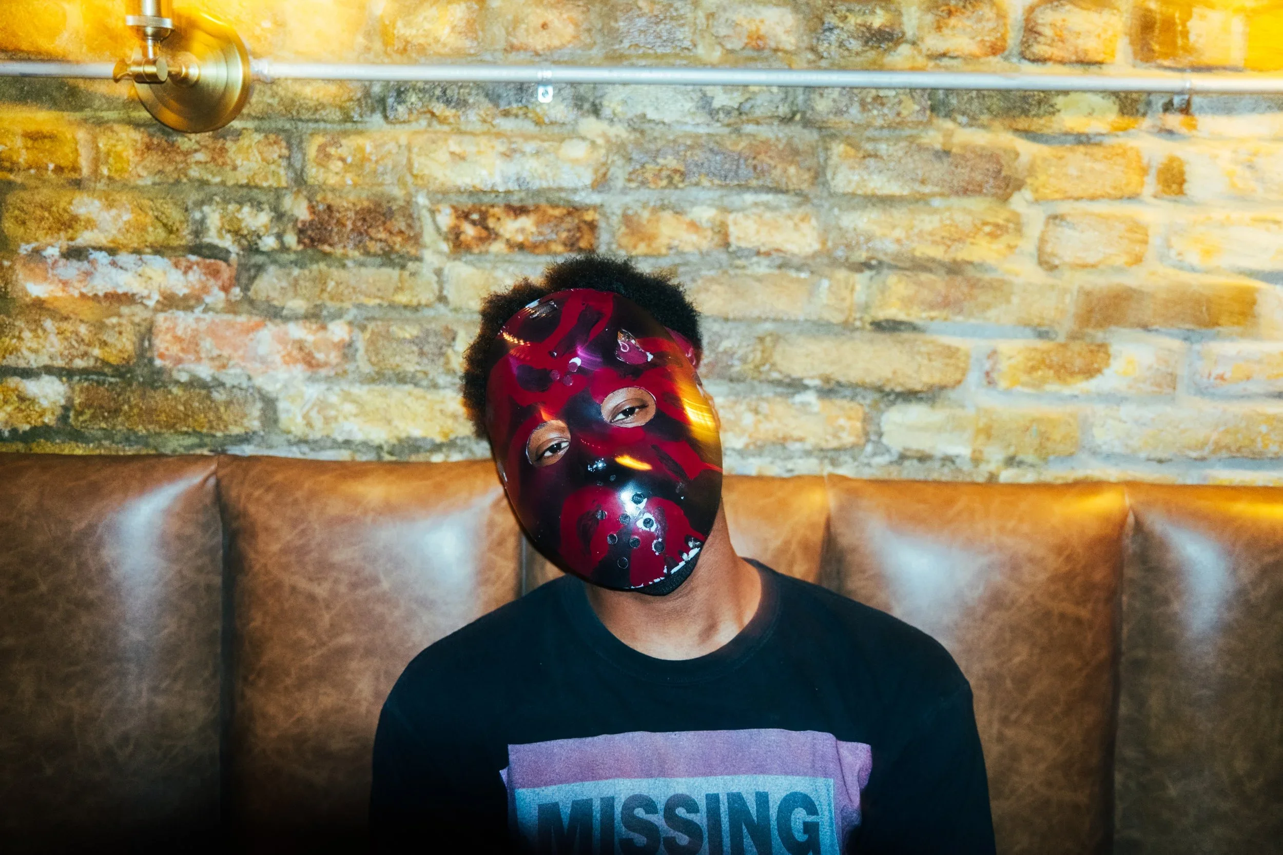 Person wearing a red and black mask with a spider design, sitting against a brick wall.