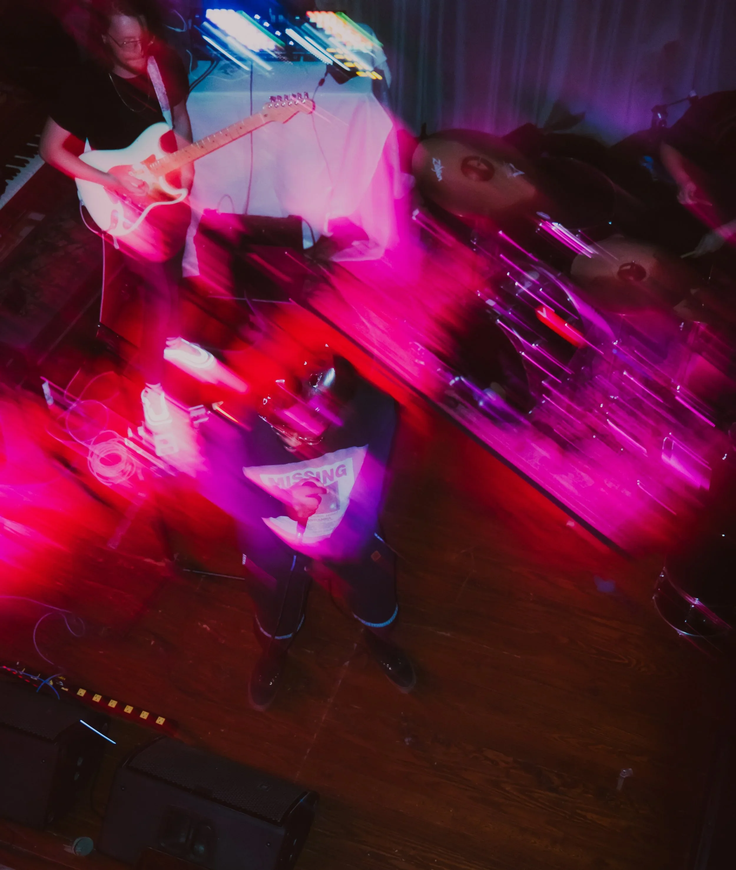 A person playing an electric guitar on stage with a digital mixing console, drum set, and synthesizer in the background, illuminated by colorful stage lights with pink and blue hues.