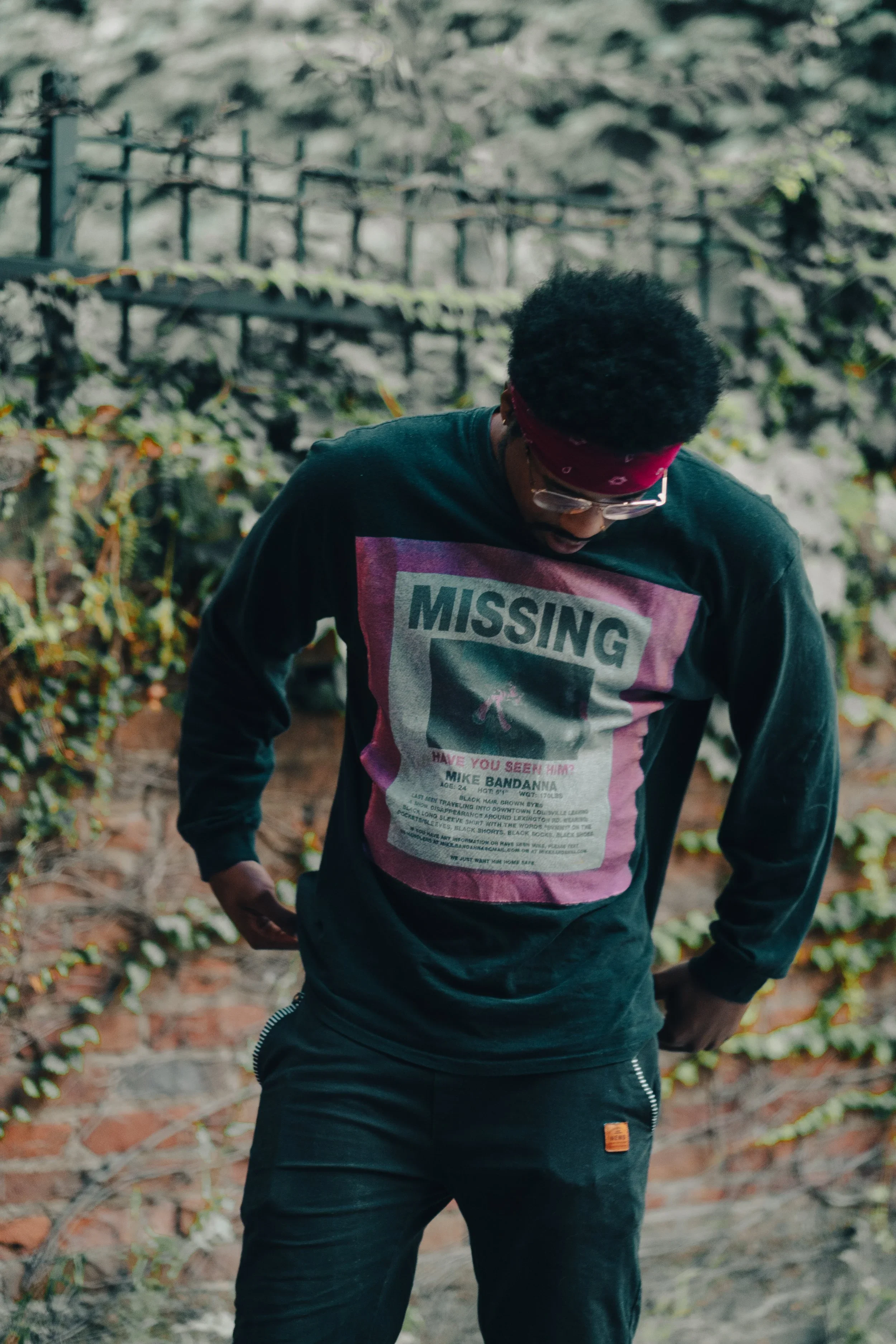 A man wearing a red bandana, glasses, black pants, and a black long-sleeve shirt with a graphic that says 'MISSING' stands outdoors near a brick wall with ivy, with his head tilted downward and hands on his hips.