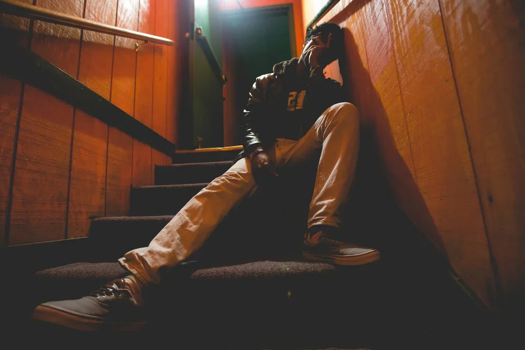 Person sitting on stairway leaning against a wooden wall, wearing a black hoodie and beige pants, with a hand on their head.