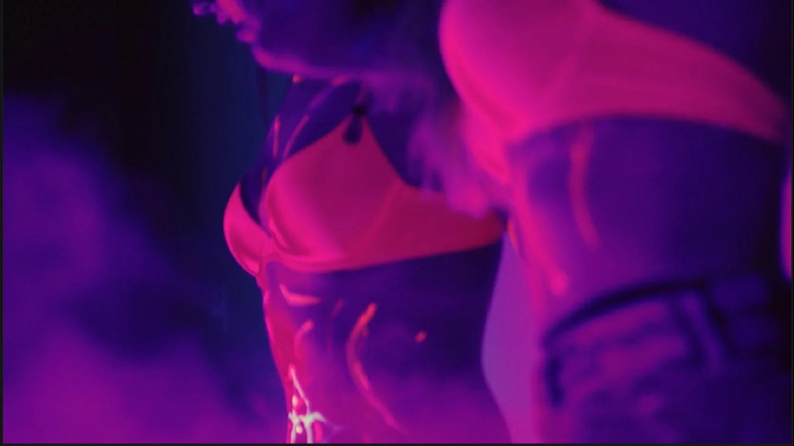 Close-up of a person's torso wearing a pink and purple jacket with a logo on the sleeve, under vibrant purple and pink lighting.