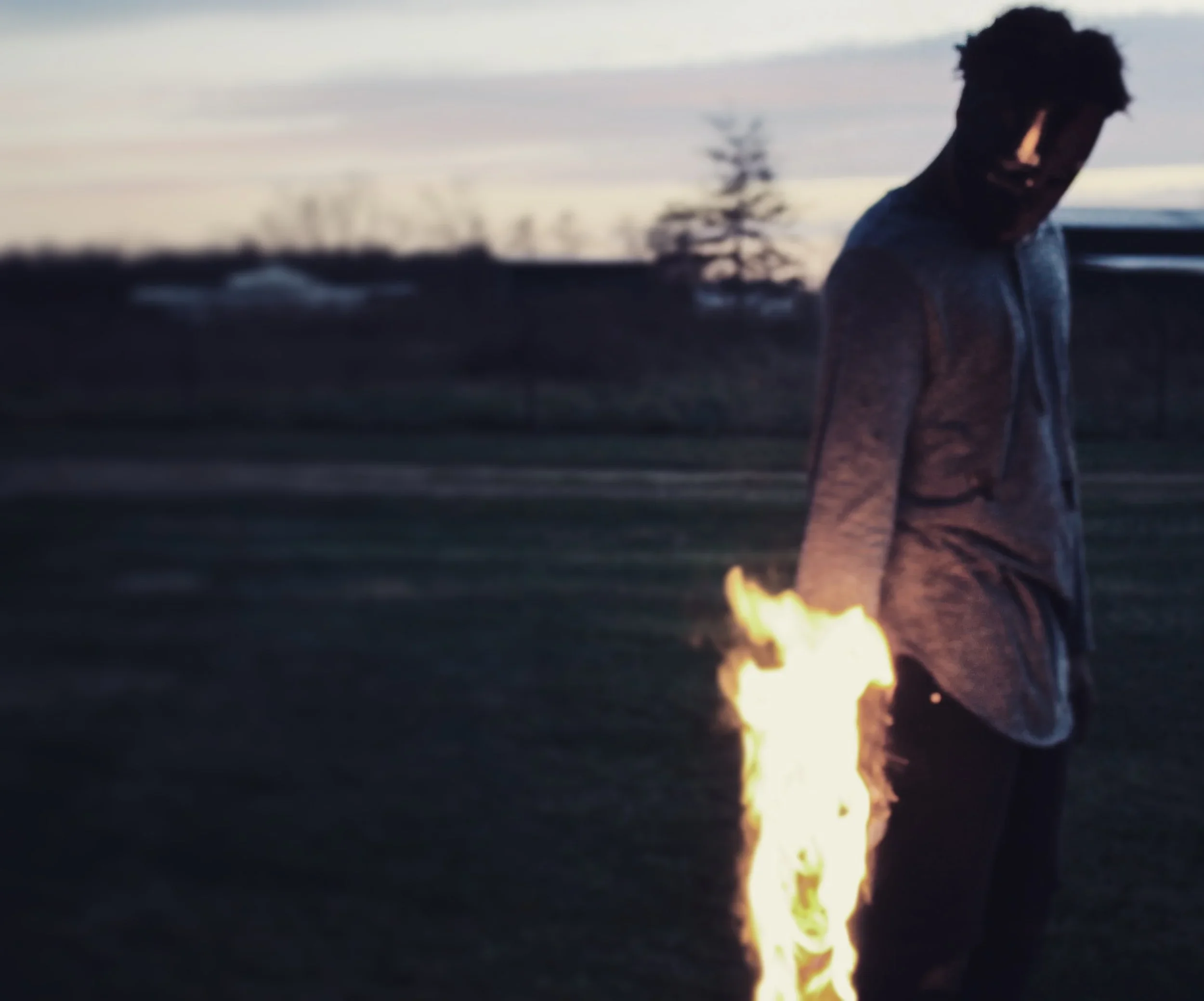 Person standing outdoors near a small fire during dusk with a sky in the background.