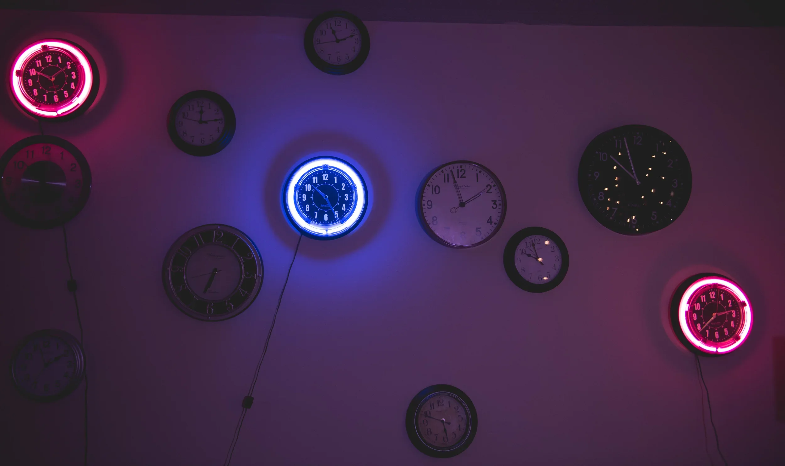 A wall decorated with multiple clocks, including neon-lit clocks in blue and pink, showing different times.
