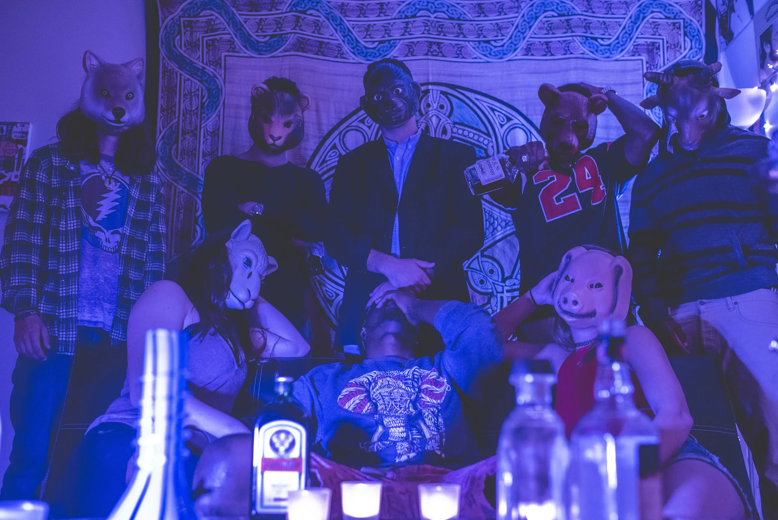 People in costumes with animal masks, including a wolf, lioness, dog, deer, and pig, posing at a party with a tribal patterned backdrop and dim lighting.