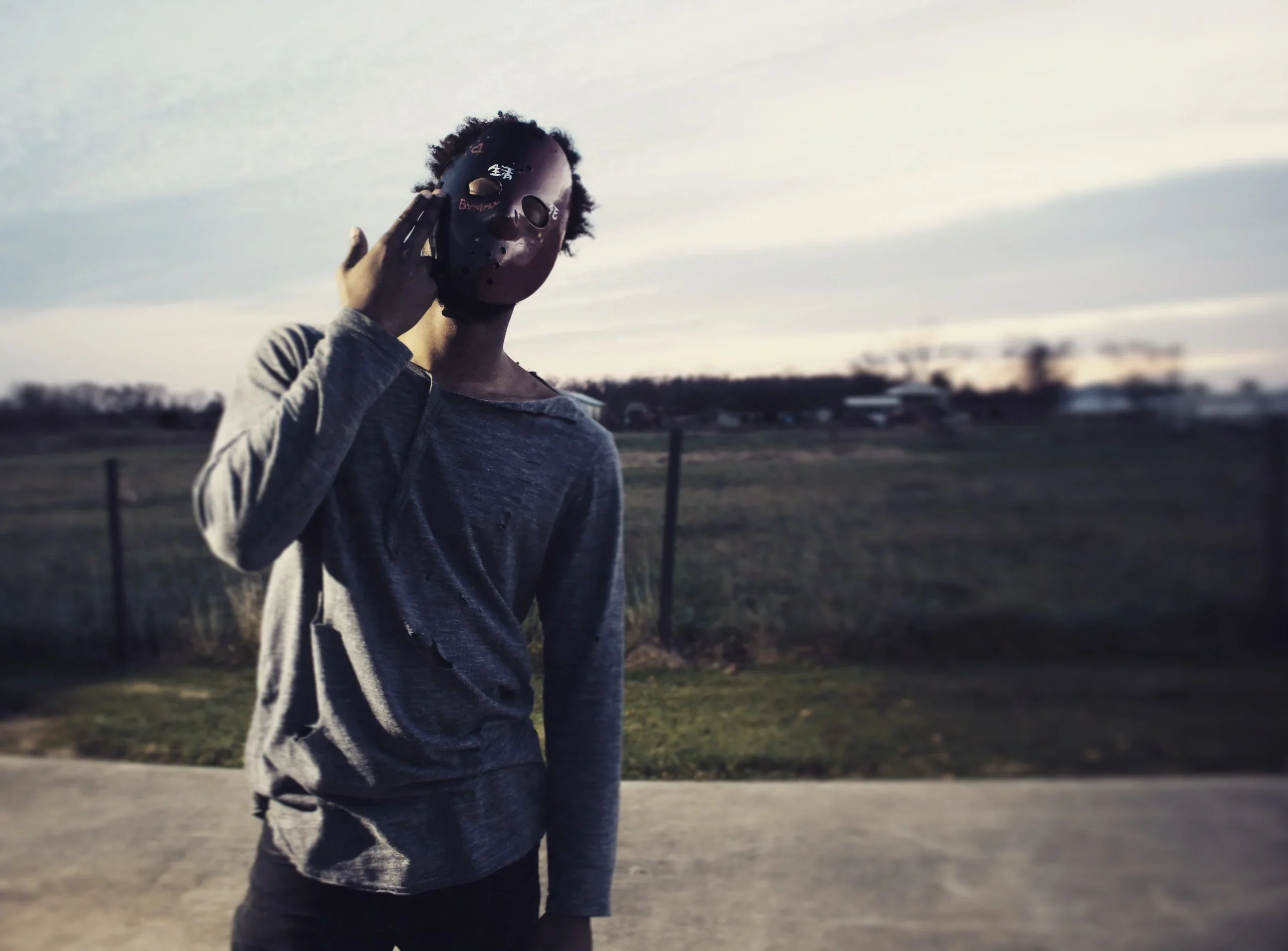 Person standing outdoors holding a black mask in front of their face during late evening or sunset.