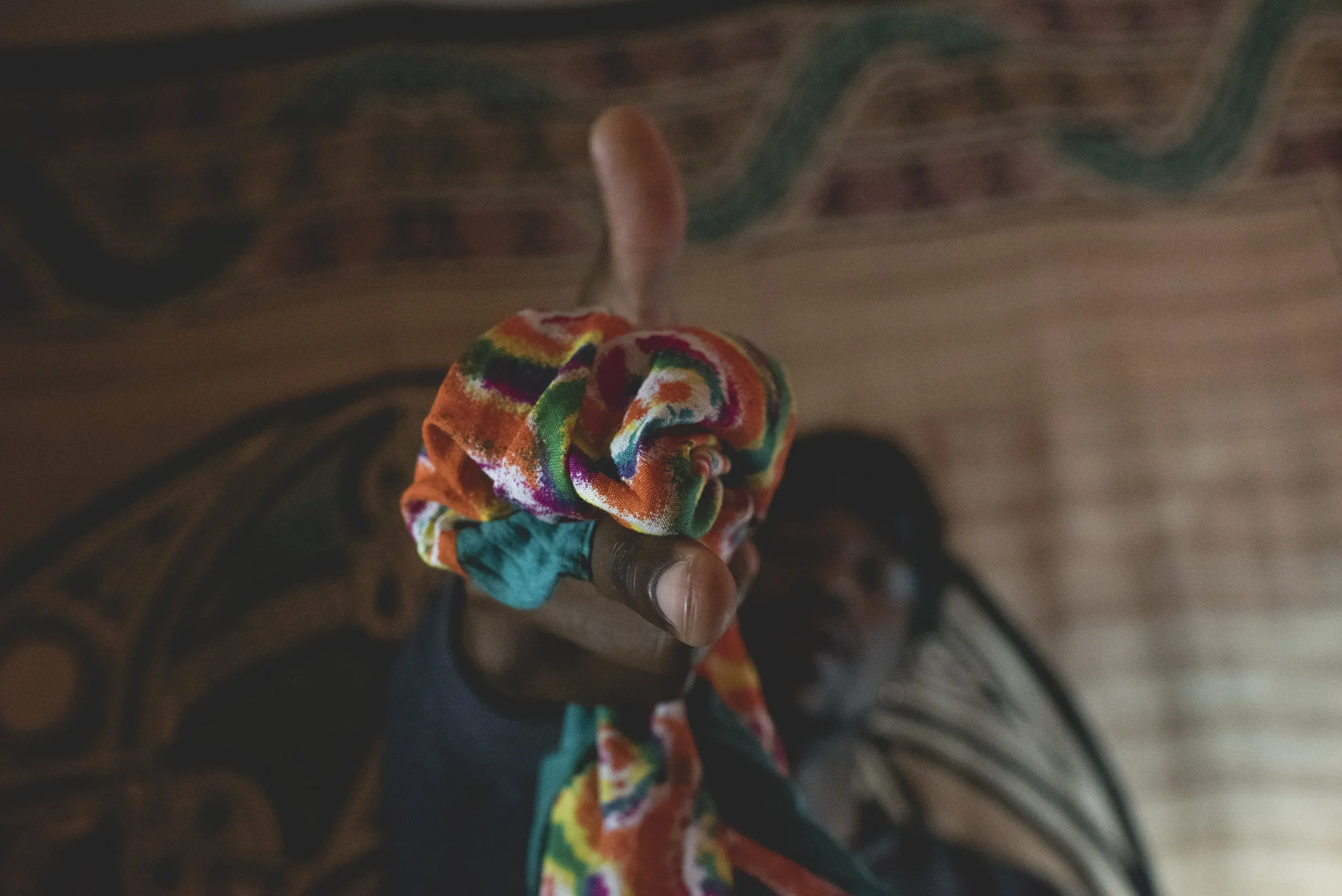 A person is holding a small, colorful, tie-dye scrunchie or fabric item with their hand, with a patterned, carpeted floor underneath.