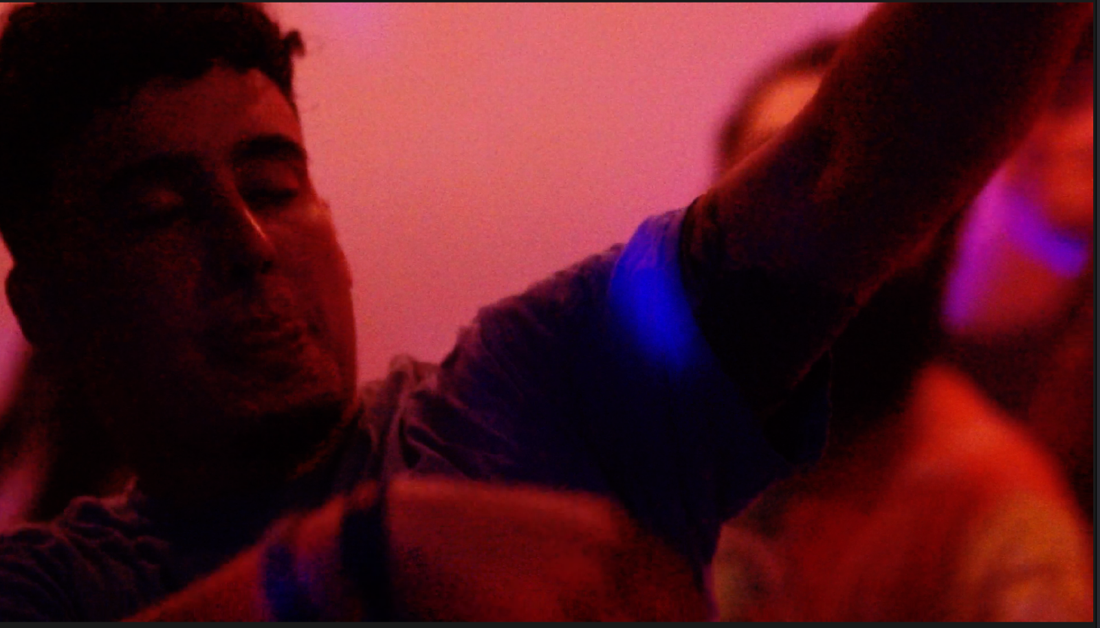 A person with closed eyes extending their arm towards the camera in a dimly lit pink and purple room.