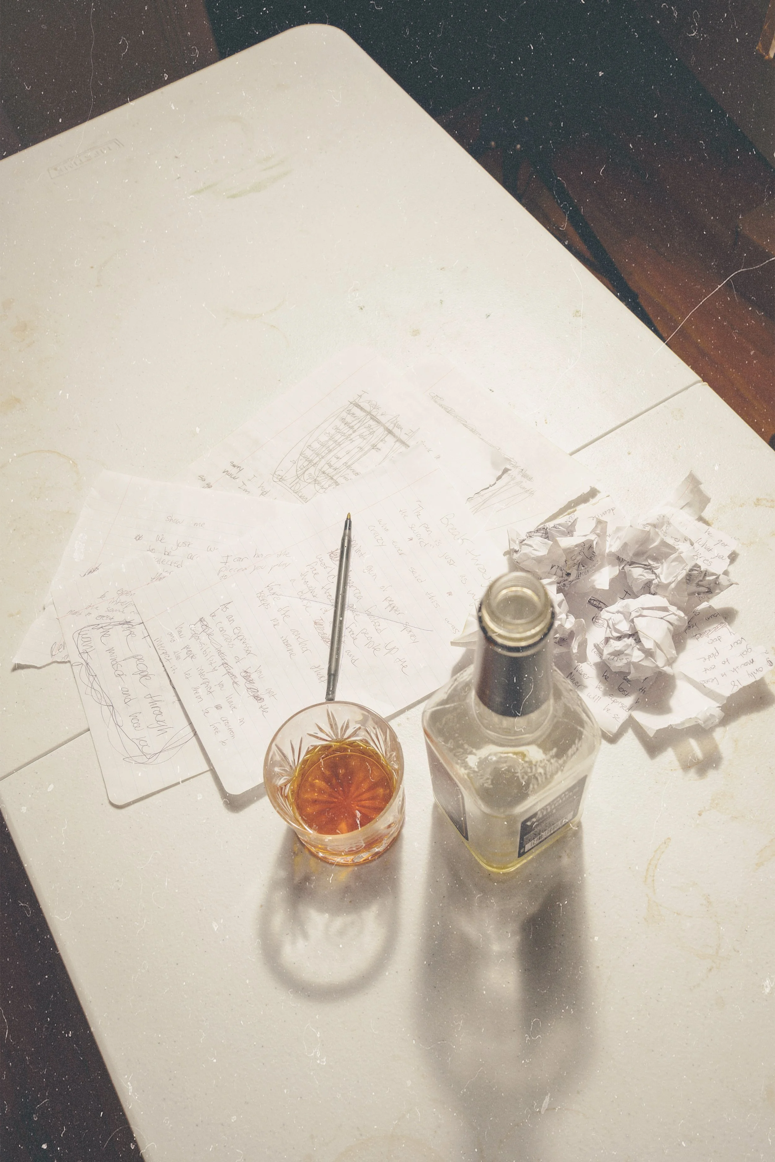 Overhead view of a white table with crumpled papers, handwritten notes on loose sheets of paper, a glass of amber-colored drink, a bottle of liquor, and a glass of water, with a dark background.