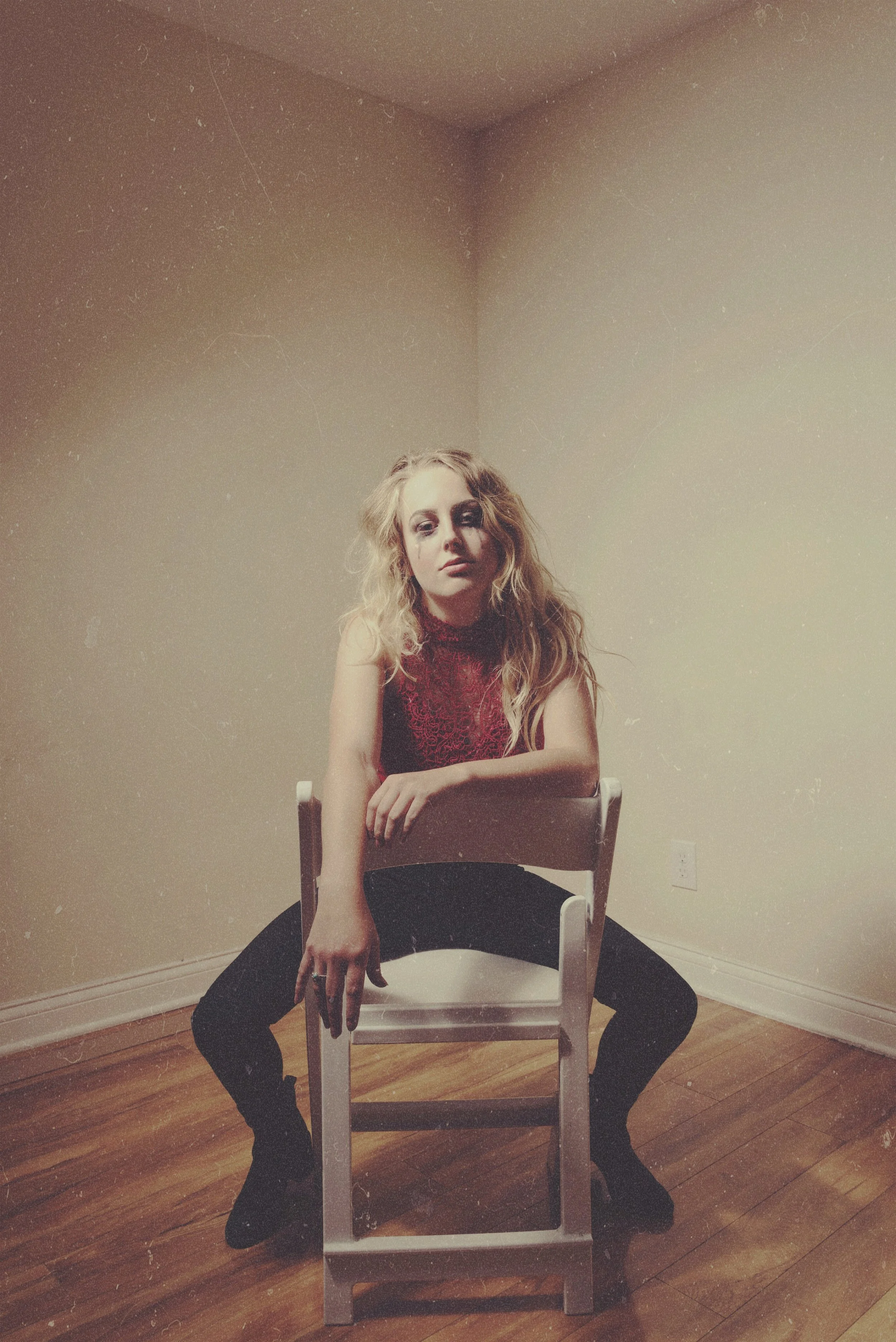 A woman with long, wavy hair sits on a white wooden chair with her legs spread apart in a room with beige walls and wooden floor. She wears a red lace top, black pants, and black boots. One arm rests on the top of the chair, and she looks directly at the camera with a serious expression.