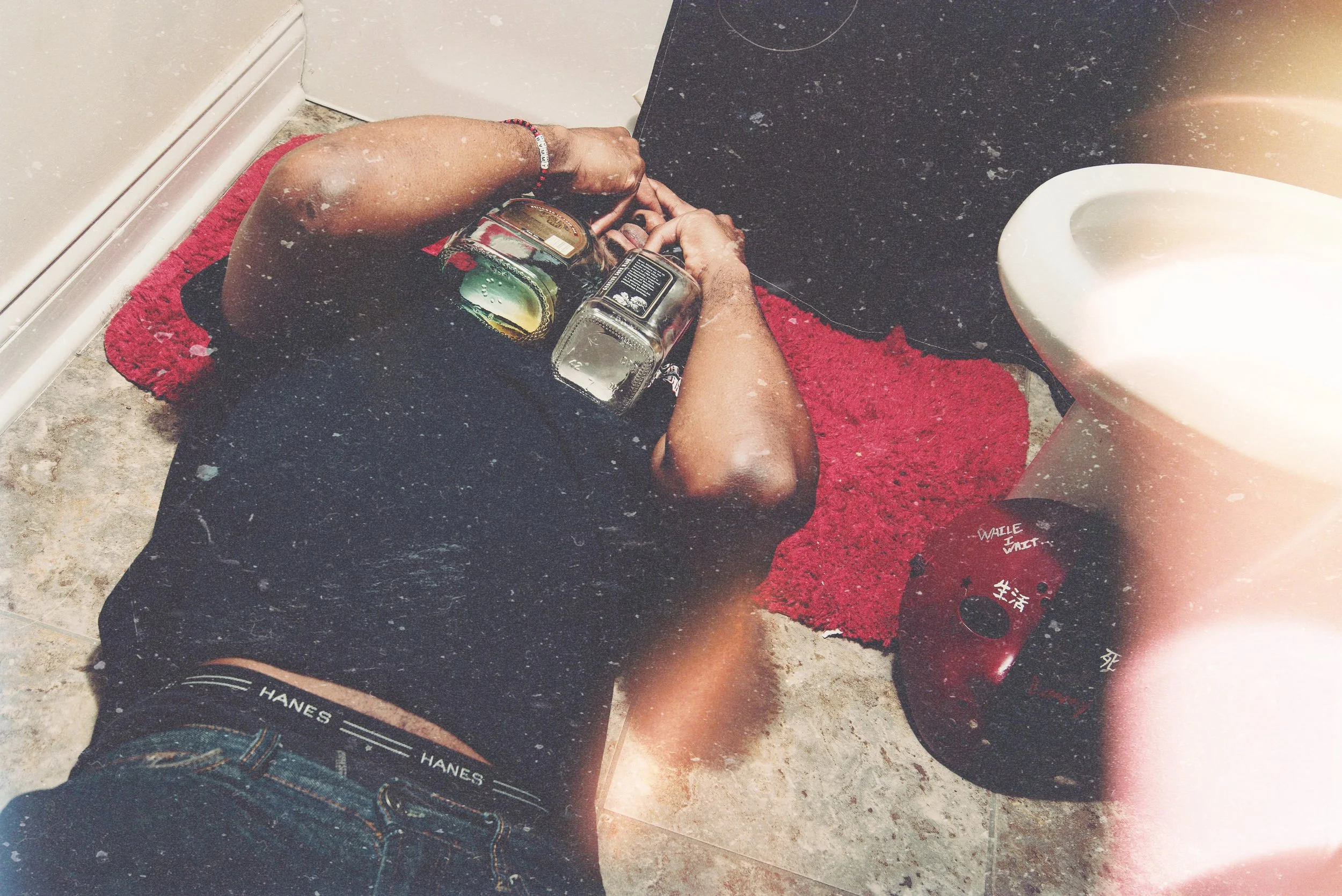 A person lying on a red rug on a bathroom floor, holding a bottle of liquor, with a glass nearby. The person is barefoot and wearing shorts and a bracelet, with a tape measure around their wrist. A toilet is visible to the right.