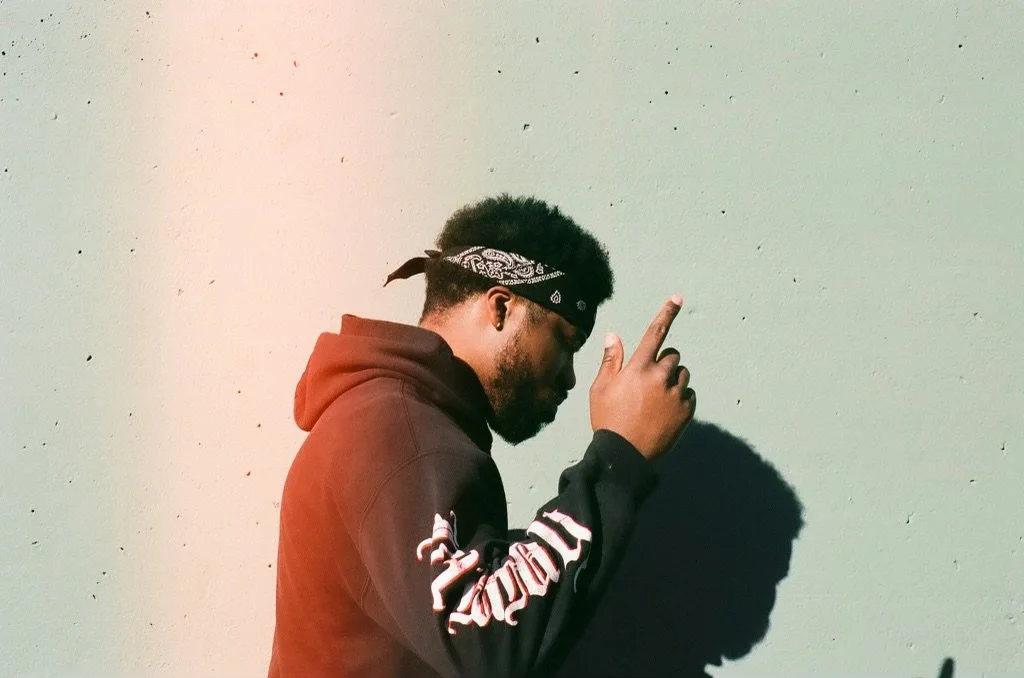 A man with a bandana on his head and a hoodie, standing against a wall, holding one finger up with his eyes closed.