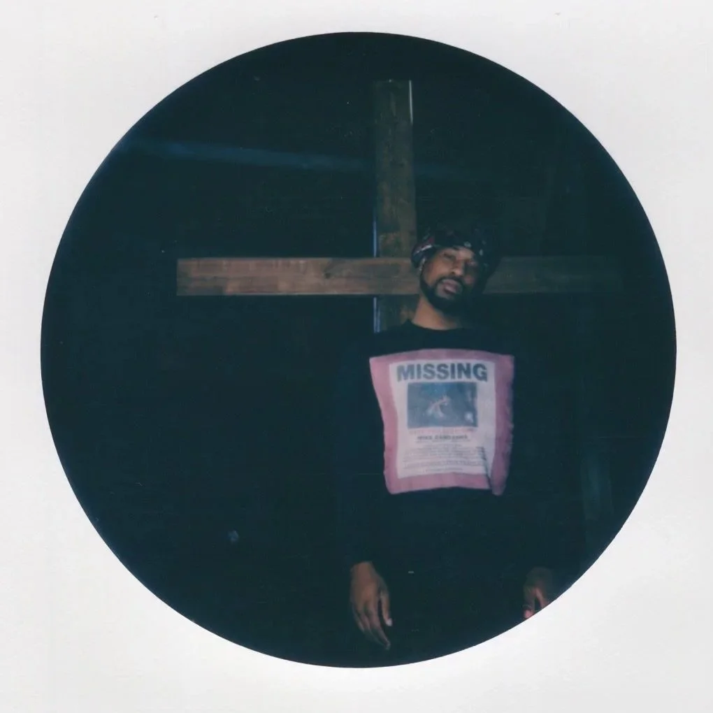 Person taking a mirror selfie, wearing a black hoodie with a picture and the word 'MISSING' on the front, standing in a dimly lit room with a wooden cross in the background.
