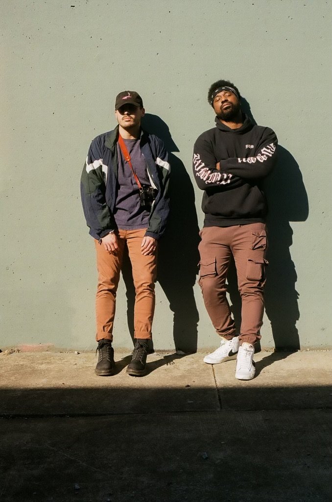 Two men standing against a plain light green wall, casting shadows, with their arms crossed or at their sides. One wears a baseball cap, sunglasses, a dark jacket, and tan pants; the other has a headband, glasses, a black hoodie, and tan cargo pants.