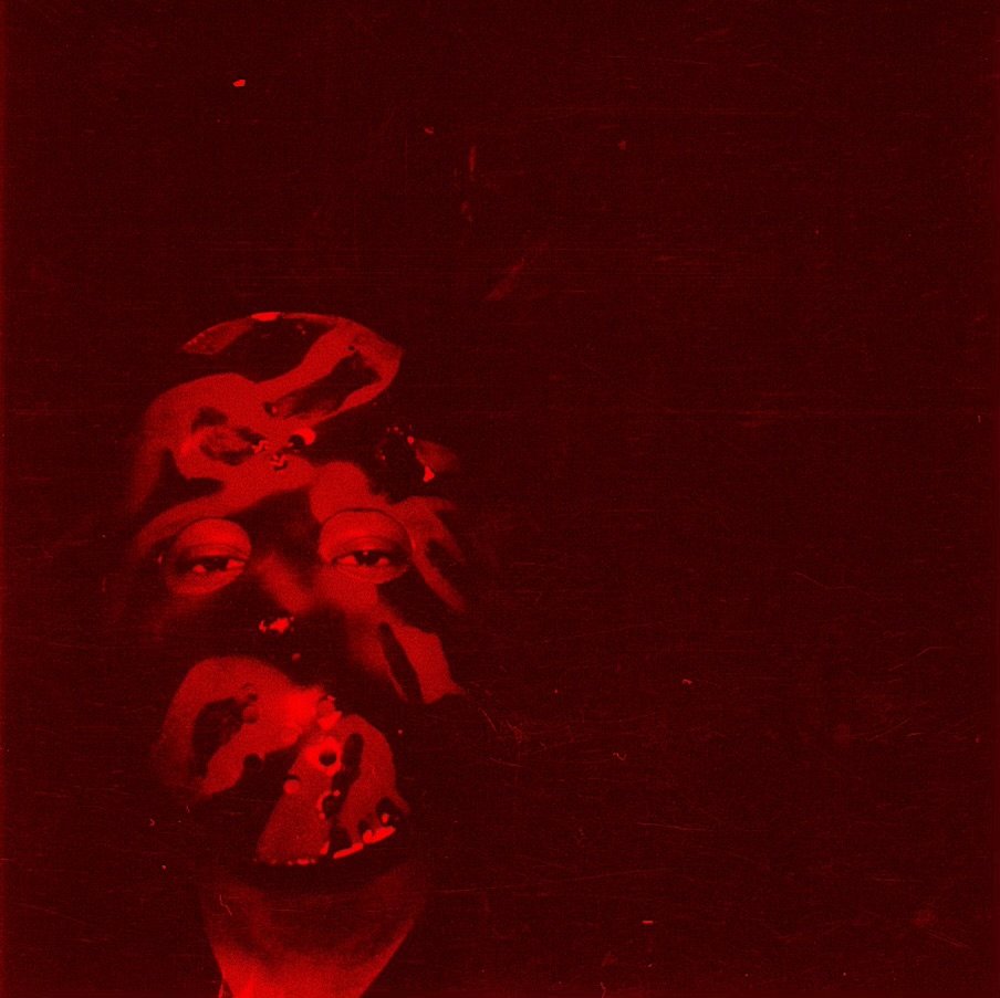 A person wearing glasses with a face painted in red and black, making a weird face with one eye and mouth open, in a dark environment.