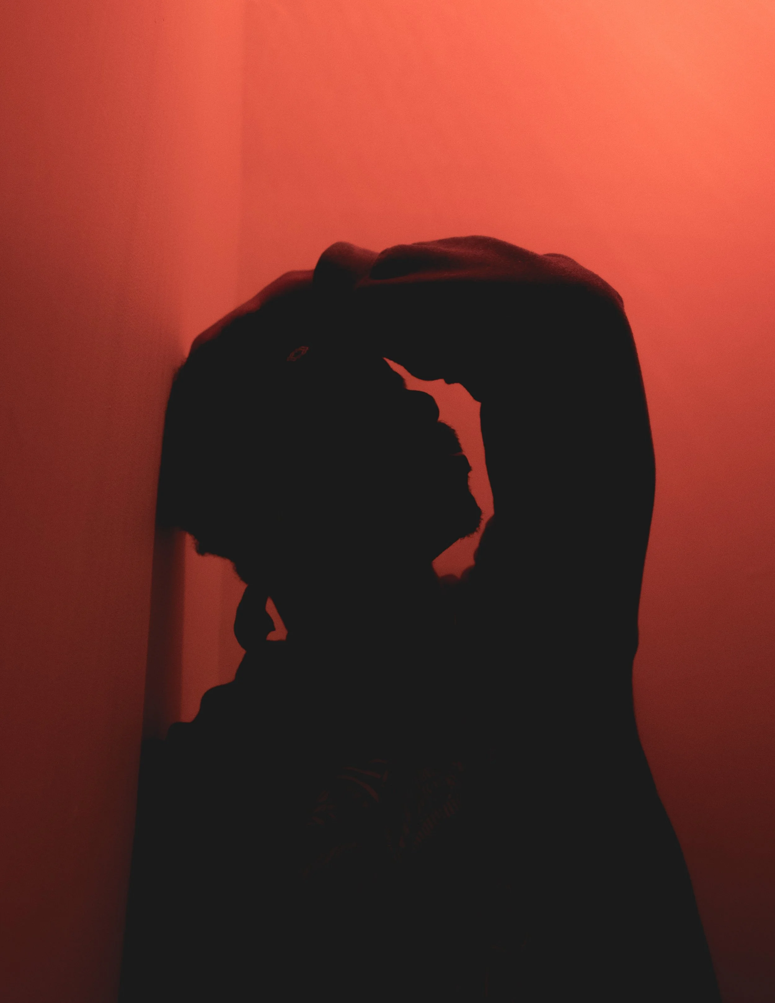 Silhouette of a person with long hair, raising their hand above their head, against a red wall background.