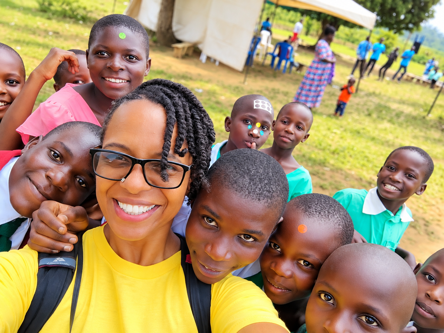A woman taking a selfie with a group of children outside on a grassy area during daytime. The woman is smiling and wearing glasses, and the children are smiling or making playful faces. In the background, there are tents, trees, and people standing or sitting.