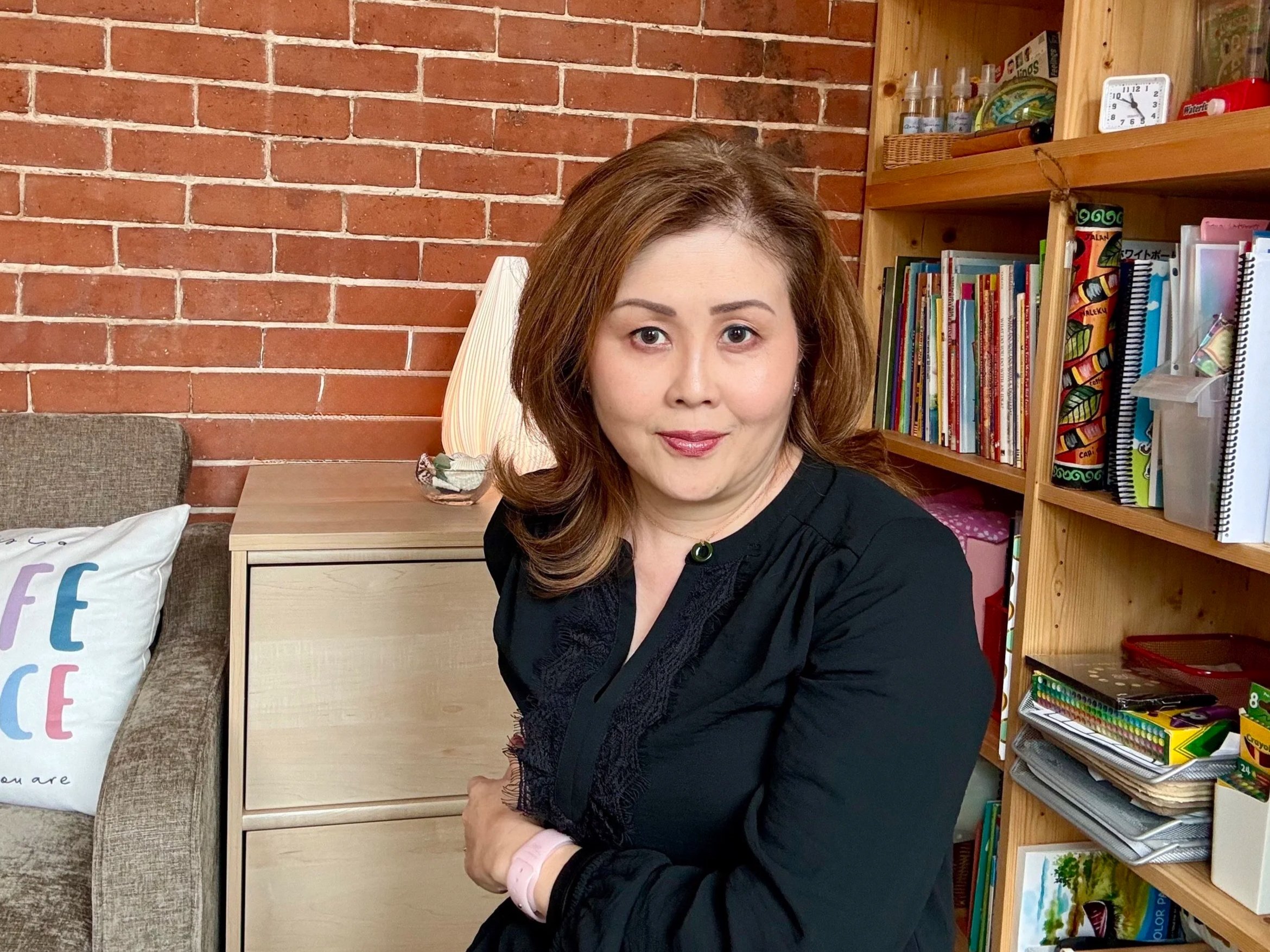 A woman sitting on a couch next to a small table with a decorative basket on top, in a room with brick walls and a bookshelf filled with books and office supplies.