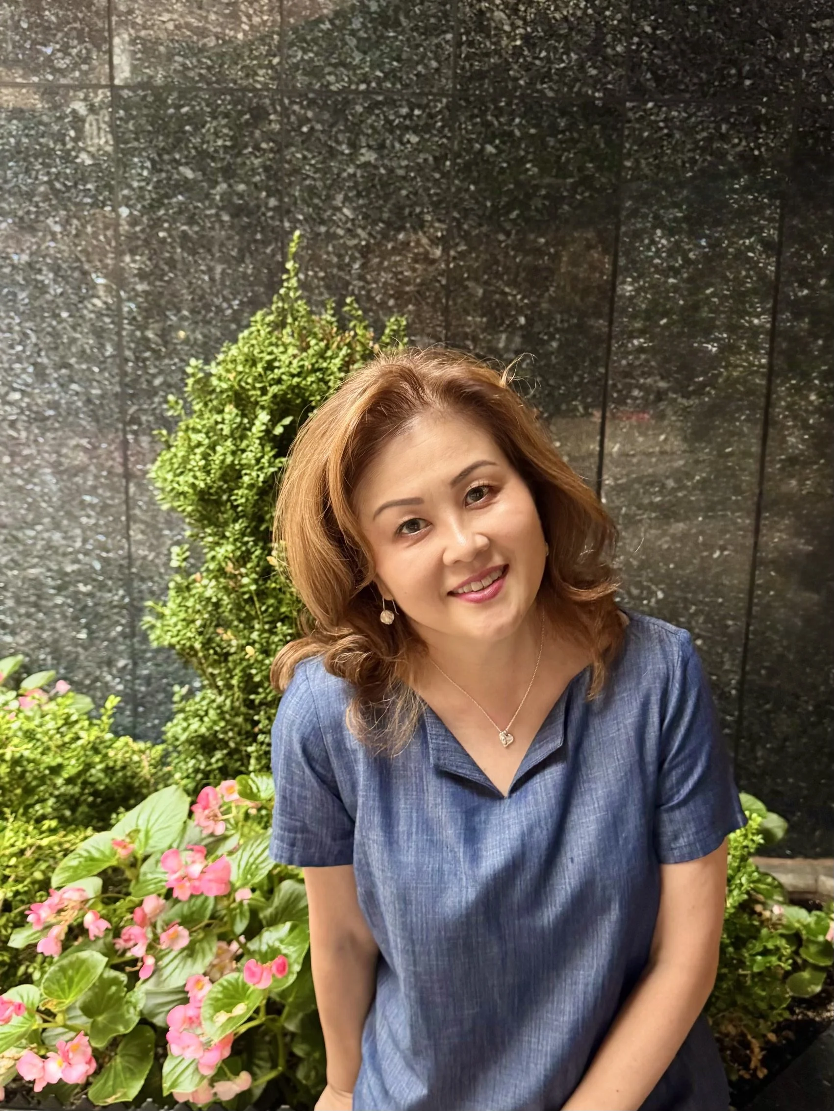 A woman with reddish-brown hair smiling, wearing a blue dress and earrings, seated outdoors with pink flowers, green foliage, and a dark textured wall behind her.