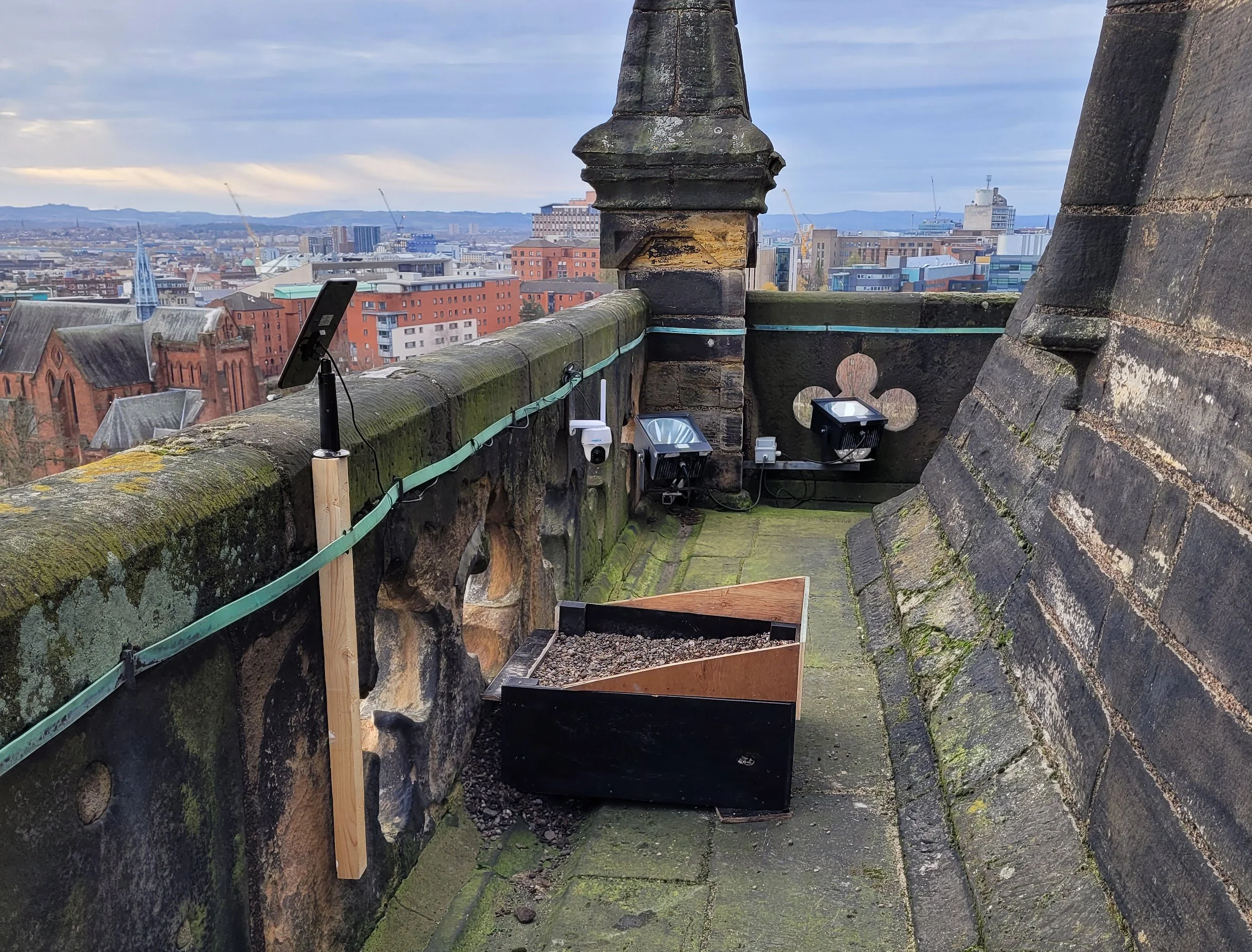 View from a rooftop or high point overlooking a city with various buildings, cranes, and a cloudy sky in the background. On the rooftop, there are equipment and safety features, including a tablet on a stand, security cameras, and solar-powered lights, with moss and weathered stonework.