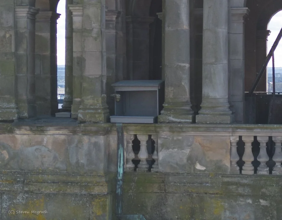 Close-up view of a historic stone building with columns and balustrades, featuring a small metal shelter on the balcony, and water dripping from the structure. The weathered stone surface shows moss and signs of aging.