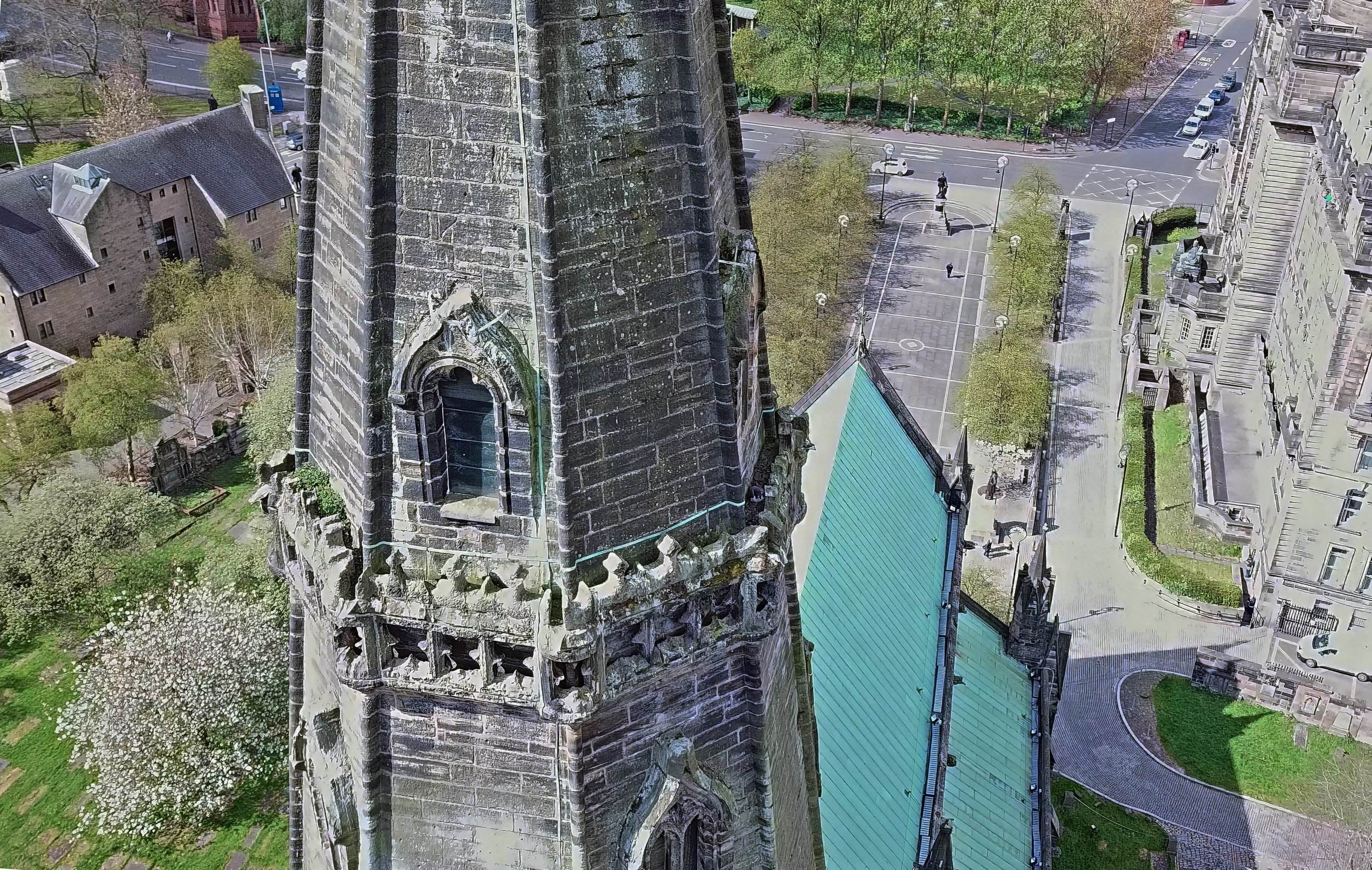 Aerial view of peregrine falcon (L7/Bonnie) on top of Glasgow cathedral with gothic architecture, surrounded by trees, sidewalk, and nearby buildings in an urban area.