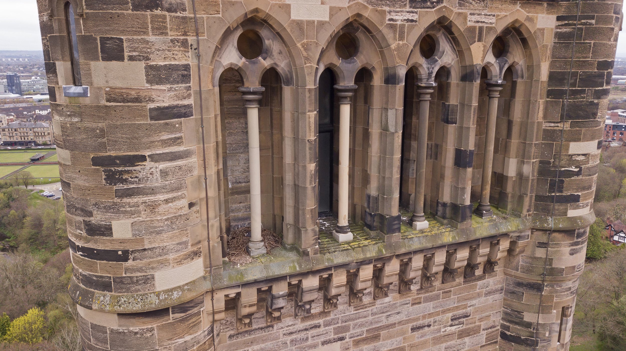 Close-up of a historic stone building with Gothic-style arched windows and columns, overlooking a cityscape and green park area.