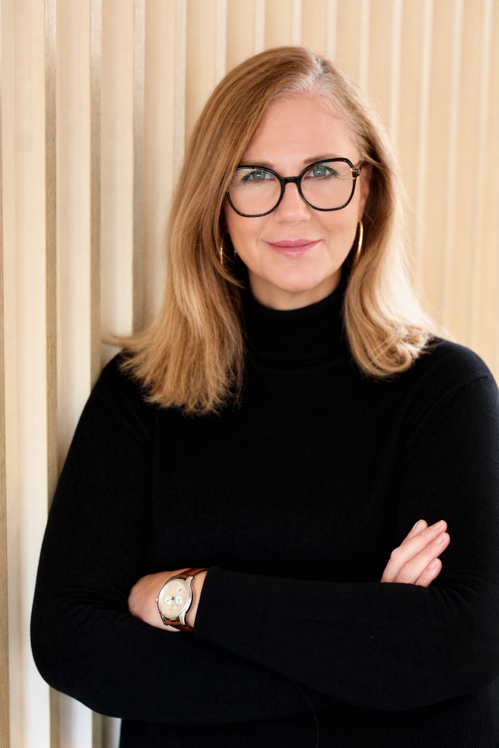 A woman with shoulder-length blond hair wearing glasses and a black turtleneck, standing with her arms crossed against a wooden background.