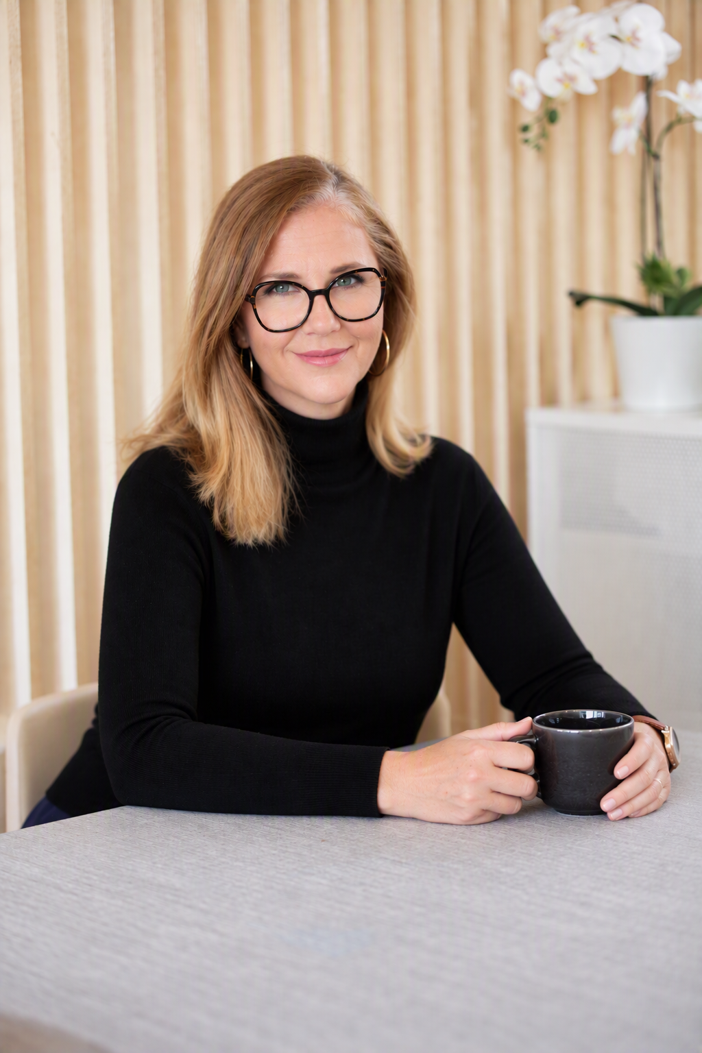 A woman with shoulder-length blonde hair, glasses, and hoop earrings sitting at a table with a black mug, wearing a black turtleneck, in a room with beige curtains and a potted orchid in the background.