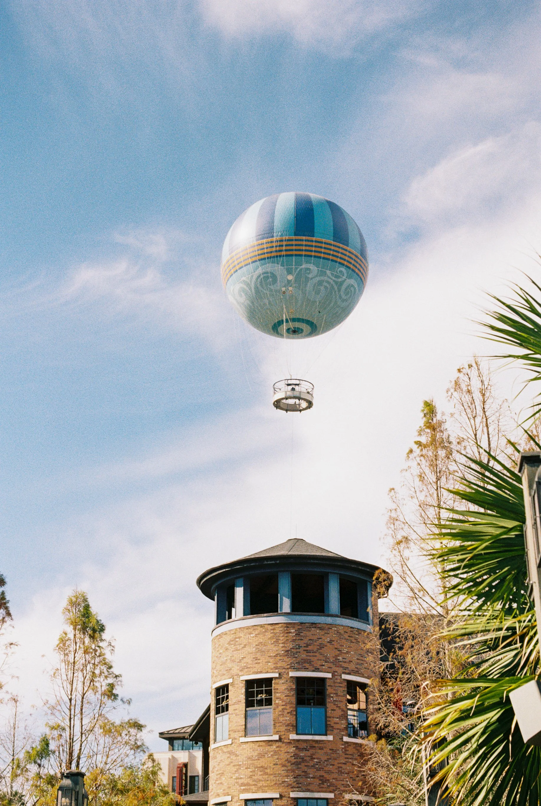 A tall, round brick building with multiple windows and a unique rooftop structure, topped by a suspended hot air balloon with blue and turquoise stripes and decorative yellow and white patterns against a partly cloudy sky with trees and plants nearby.