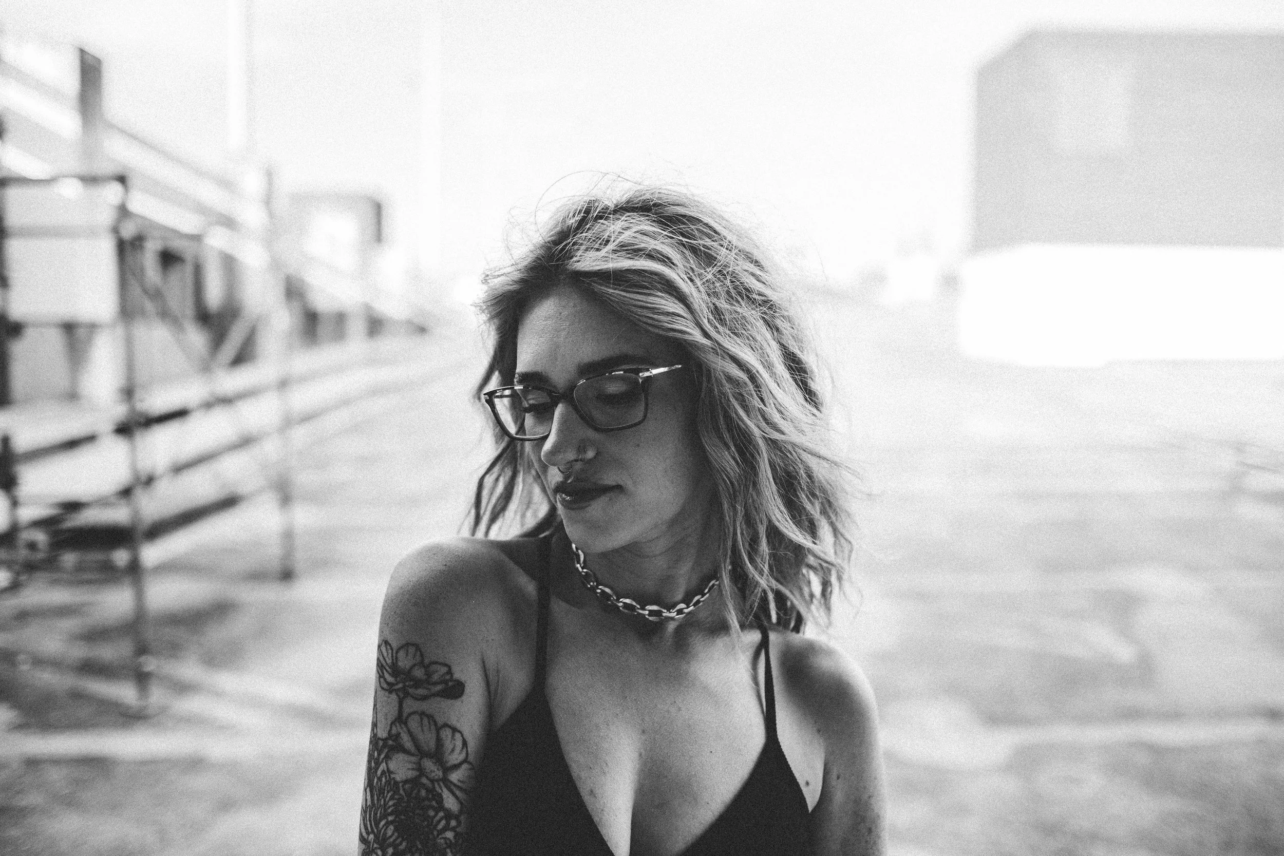 A black-and-white photo of a woman with wavy hair, wearing glasses, a chain necklace, and a sleeveless top. She has tattoos on her arm and looks downward. The background features outdoor staircase and blurred buildings.
