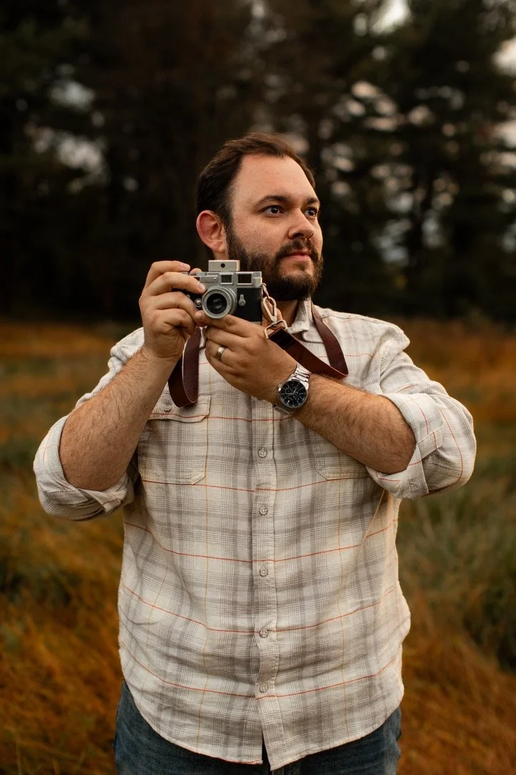 A man with a beard and plaid shirt holding a vintage camera outdoors in a natural setting.
