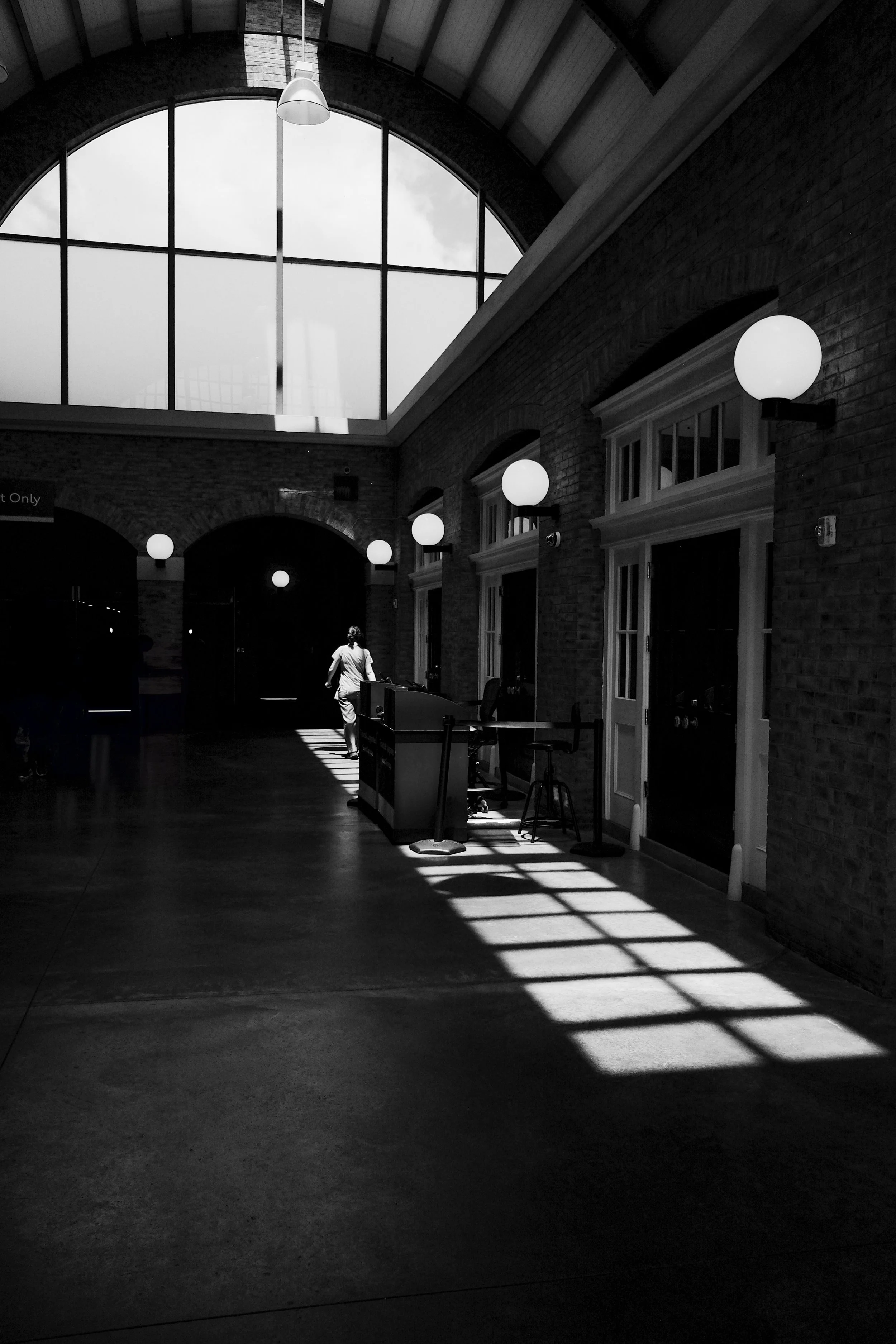 Black and white photo of an indoor space with large arched window on the ceiling, glowing spherical wall lights on brick walls, and a person walking away carrying a bag, casting shadows on the floor.