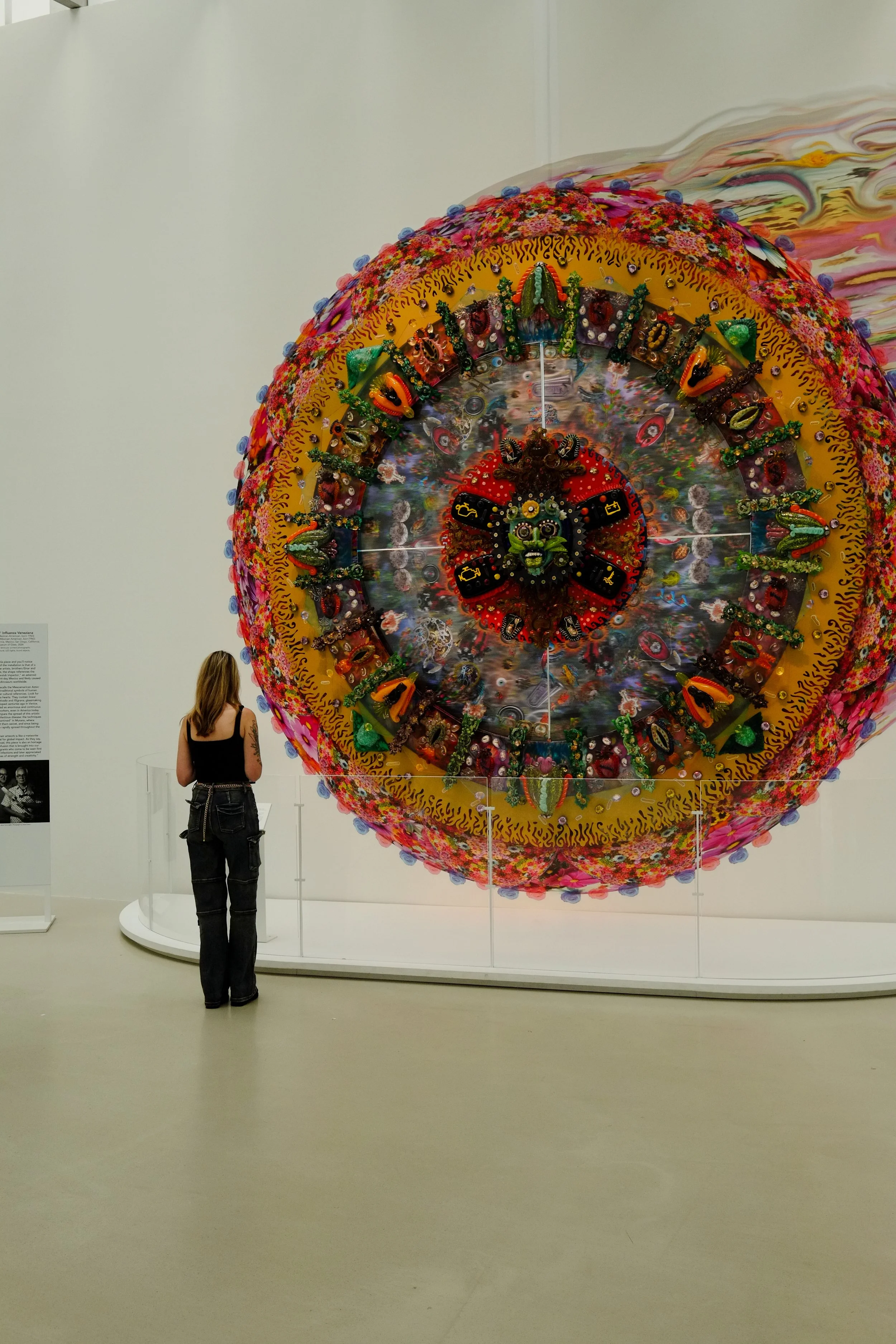 A woman with tattoos wearing a black top and jeans standing in front of a colorful, large, circular art installation inside a museum or gallery.