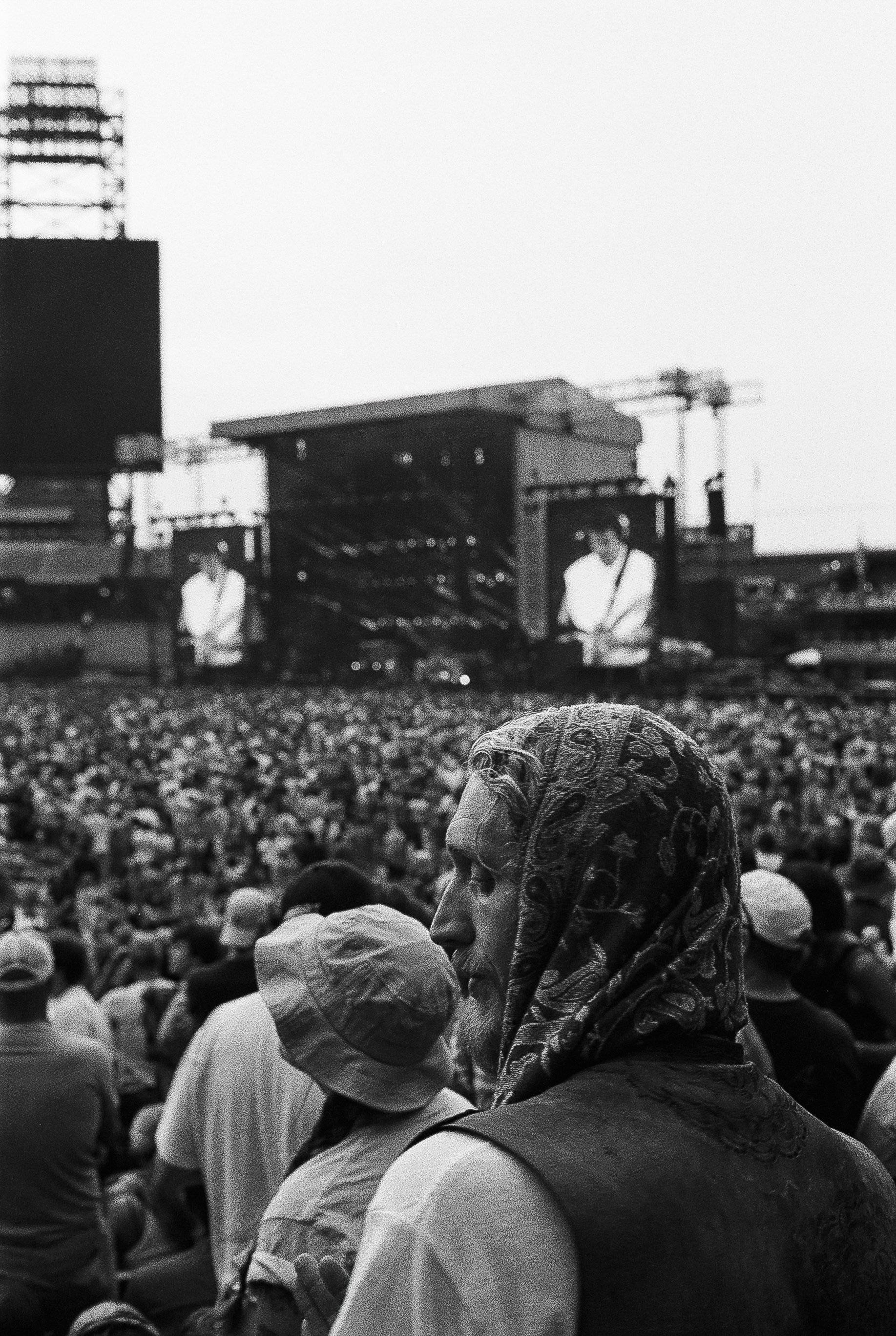 A woman wearing a headscarf facing left among a crowd of people at an outdoor event, with a stage and construction cranes in the background.