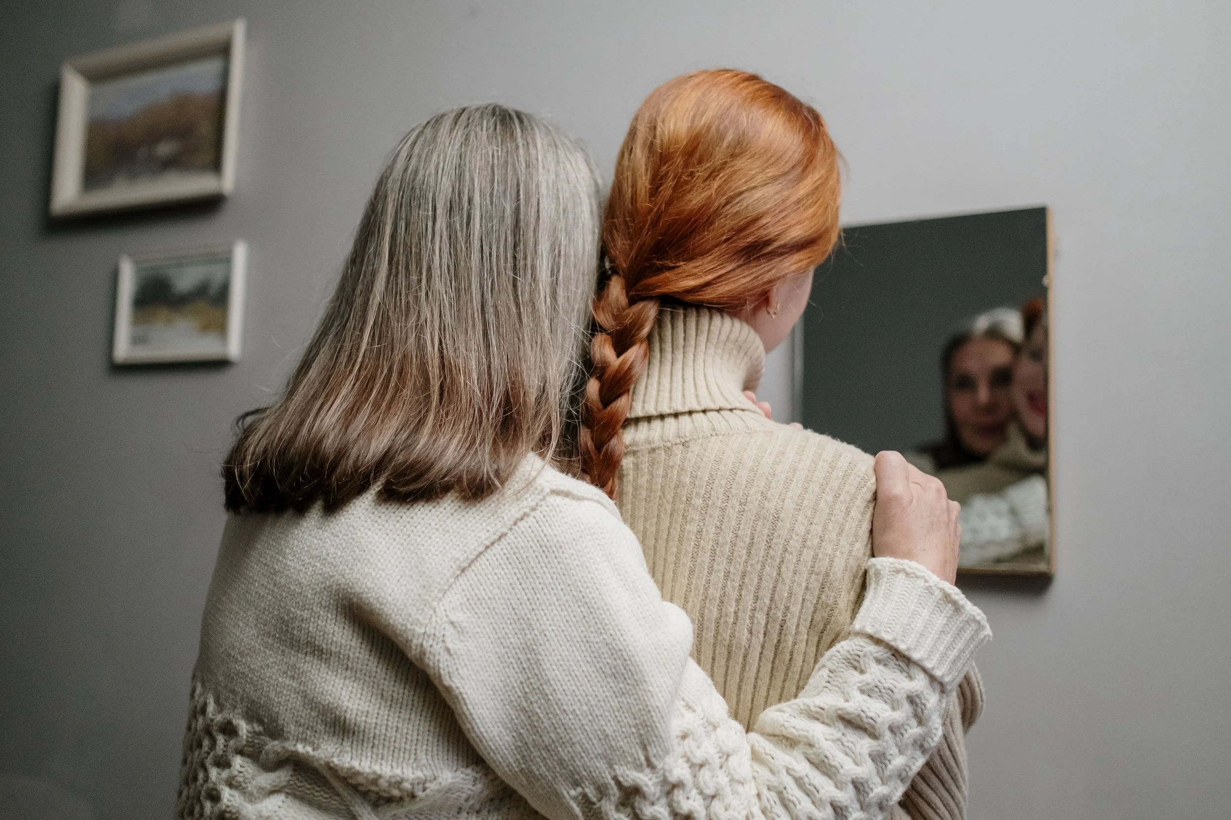 An older woman sharing a hug with the younger version of herself in front of a mirror in a room, with framed pictures on the wall.