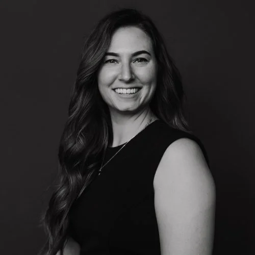 Black and white portrait of a smiling woman with long wavy hair, wearing a sleeveless top, against a dark background.