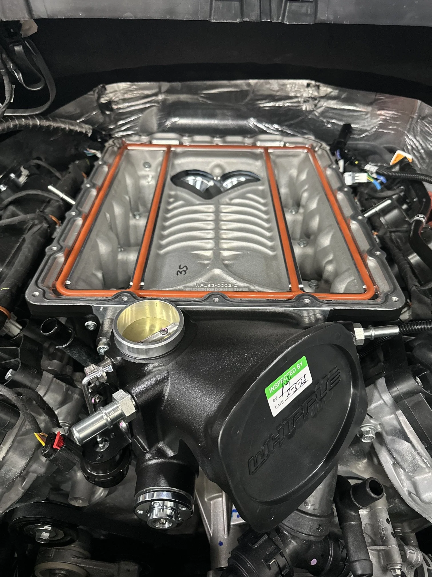 Close-up of a car engine with an orange gasket around the intake manifold, surrounded by various engine components and wires.