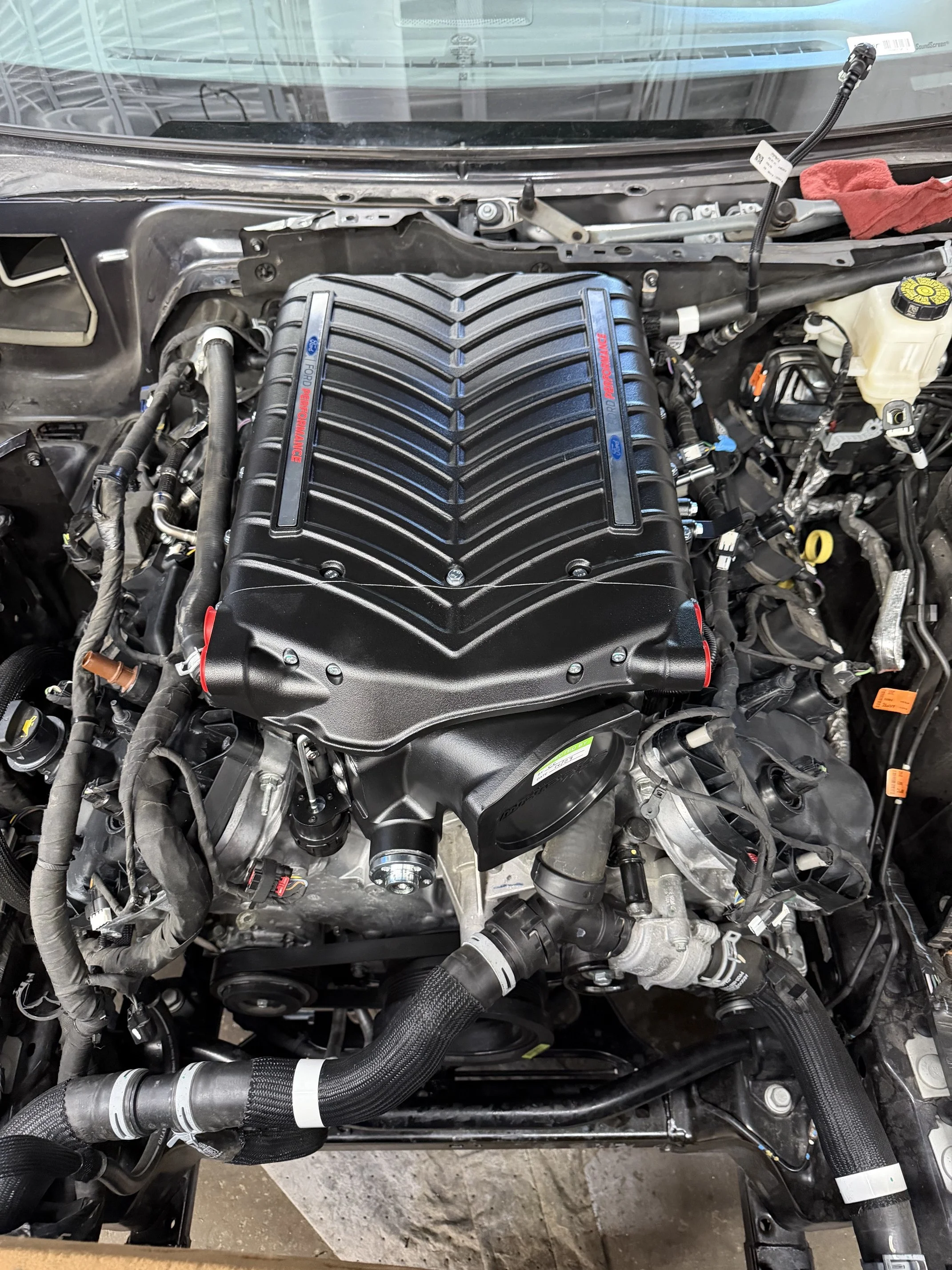 Close-up of a car engine with a black performance supercharger cover and mechanical components surrounding it.