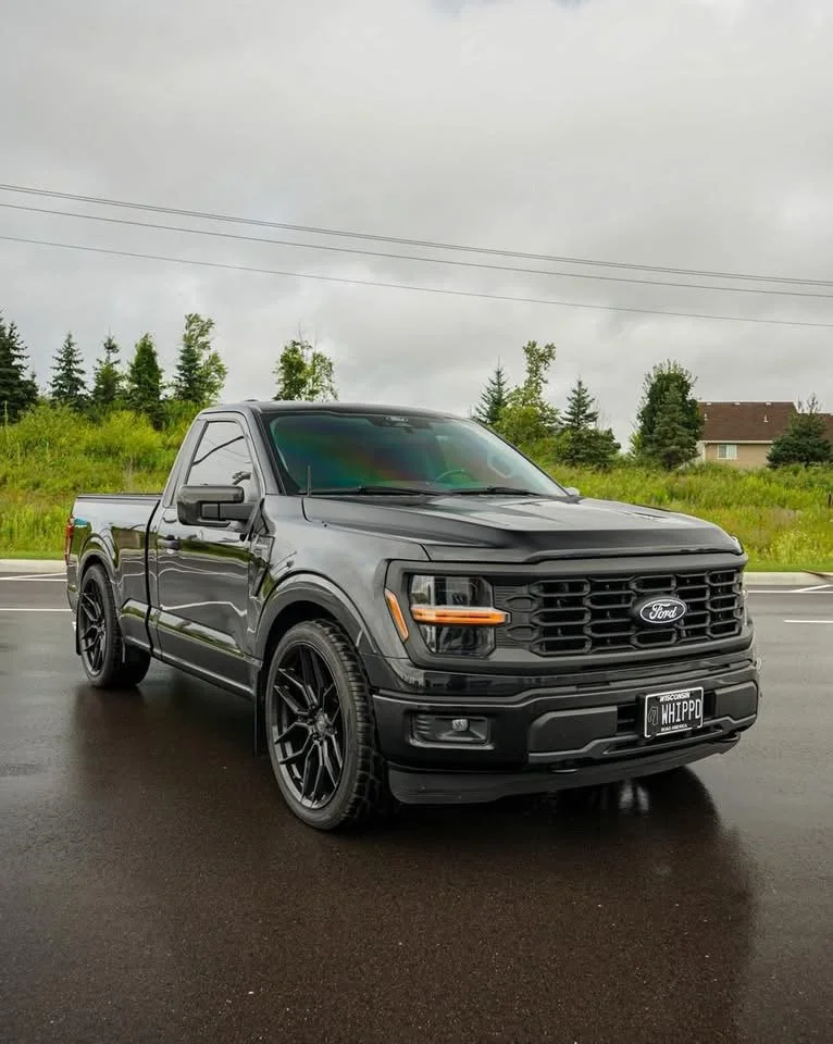 A black Ford pickup truck parked on a wet road under an overcast sky, with green grass and trees in the background.