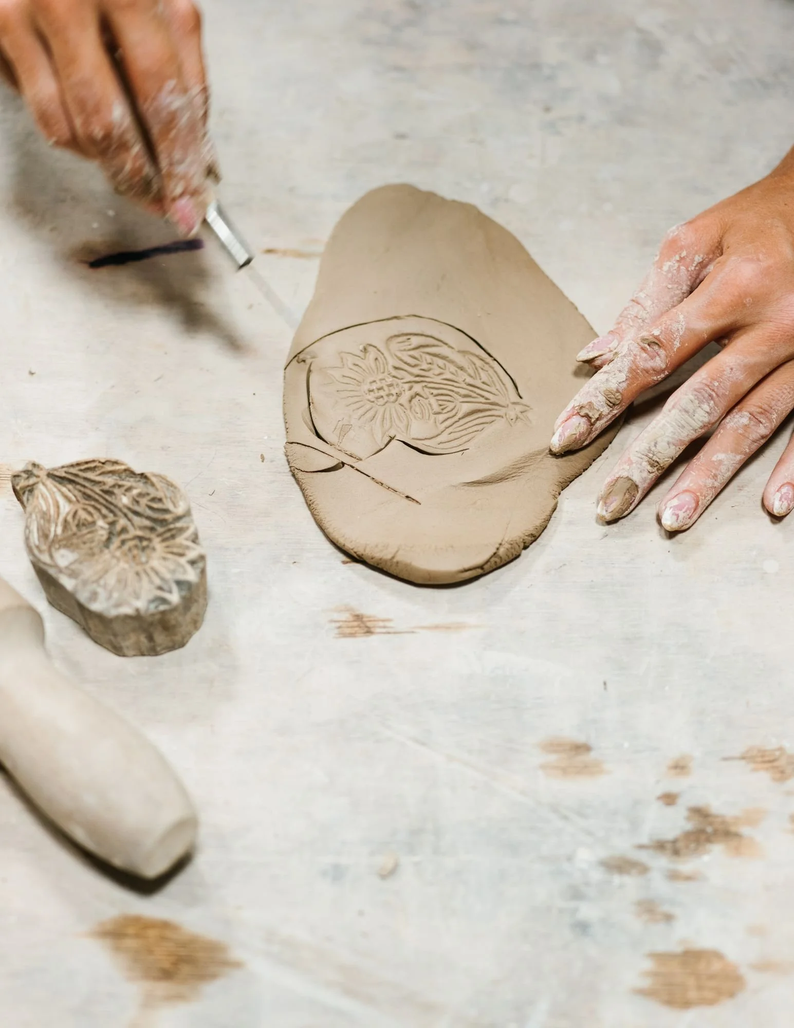Hands carving a floral design into clay with a carving tool.