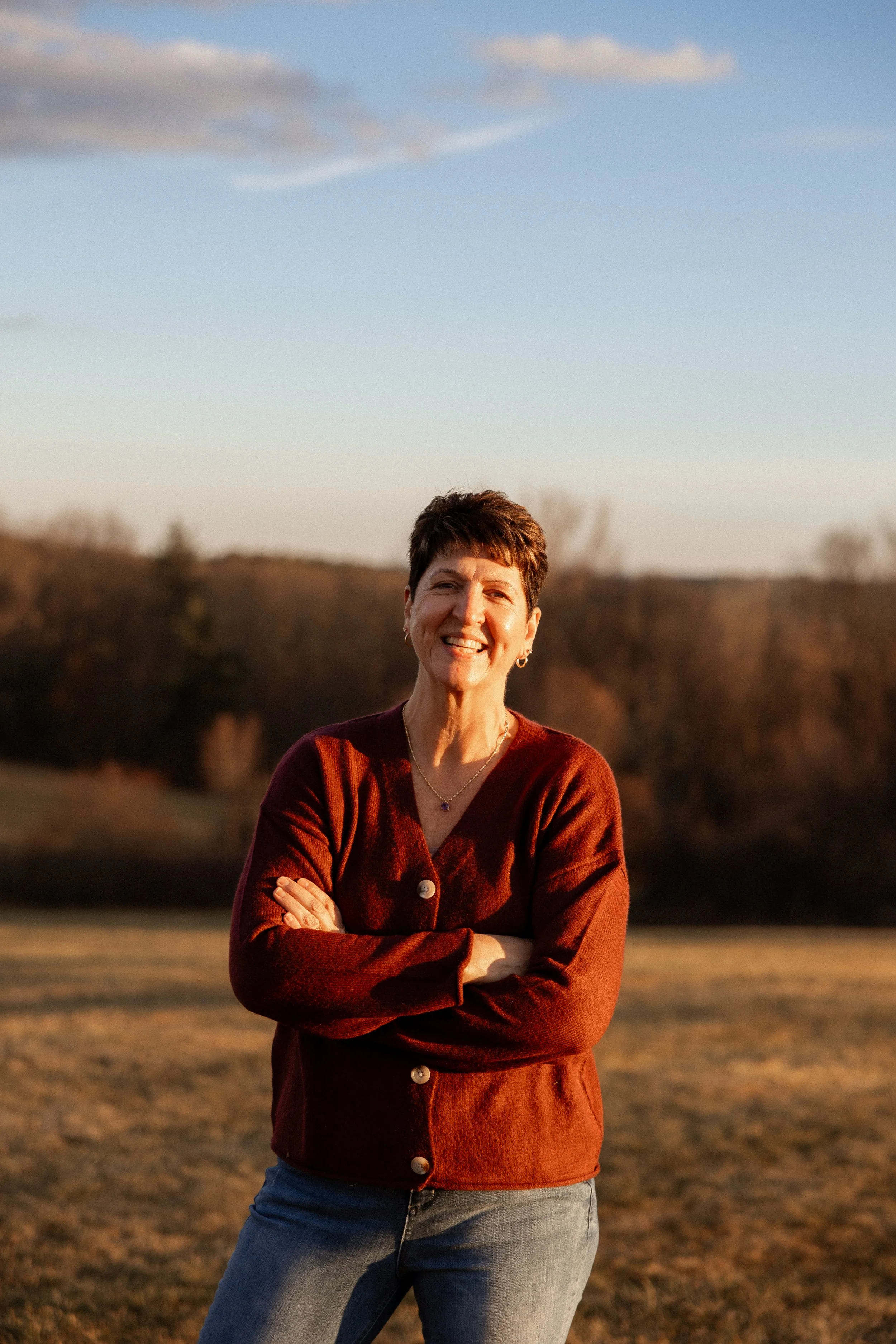 A woman smiling outdoors during sunset, wearing a maroon cardigan with buttons and blue jeans, standing with her arms crossed, in a natural landscape with trees and open sky.