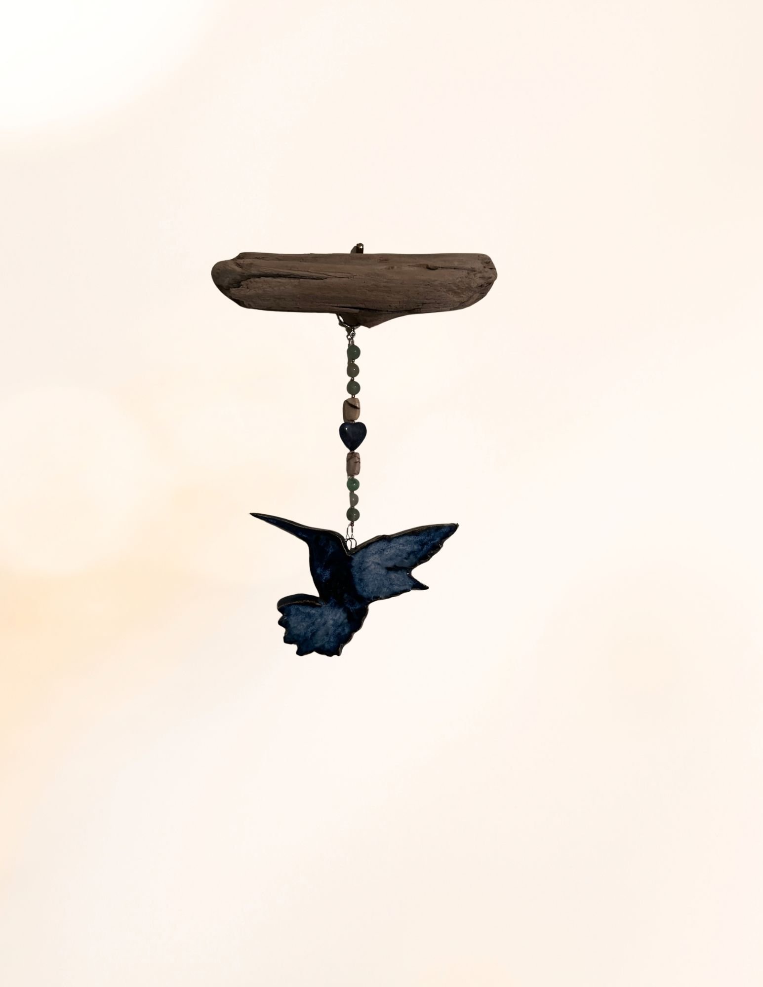 Silhouette of a bird in flight under a hanging mobile with beads and a driftwood piece against a plain background.