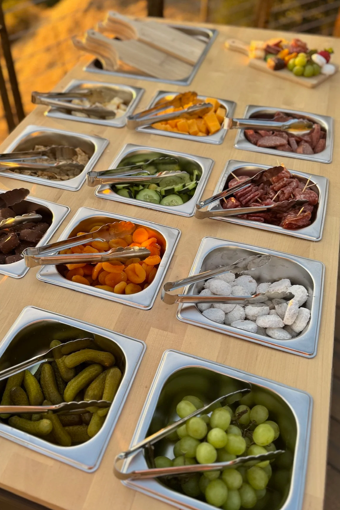 Variety of condiments and toppings for a buffet, including pickles, grapes, dried fruit, cubed cheese, sliced cucumbers, sliced meats, grapes, and assorted vegetables, with tongs in each container. The table is outdoors with a blurred background.