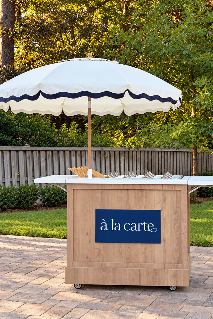 Outdoor buffet stand with an umbrella, set up on a patio with grass, bushes, trees, and a wooden fence in the background, labeled 'à la carte'.