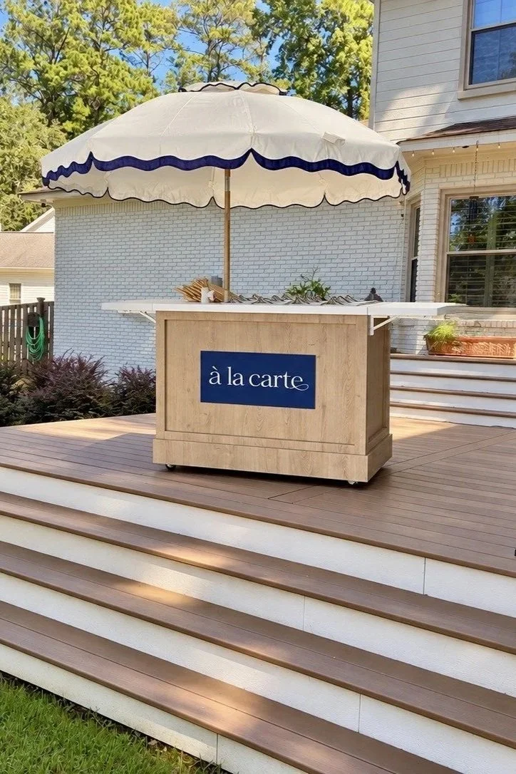 Outdoor wooden deck with stairs, a beige cart with a blue sign saying 'à la carte' underneath a large white umbrella, and a black outdoor chair. The deck is attached to a white house with large windows, surrounded by greenery and a blue sky.