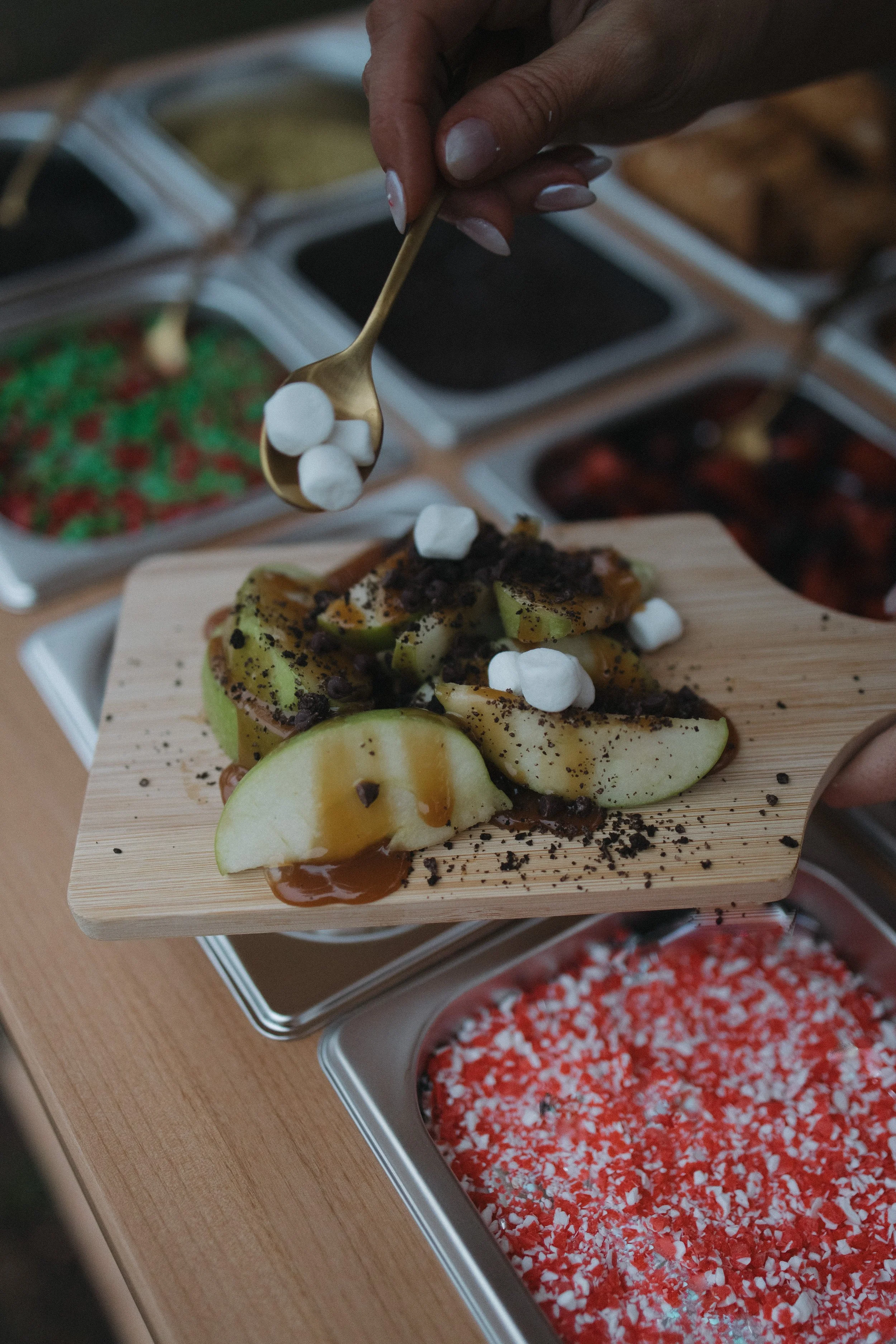 A hand holding a small gold spoon with marshmallows, above a dessert with sliced pears, chocolate crumbs, and caramel sauce, on a wooden board; background shows containers of various toppings.