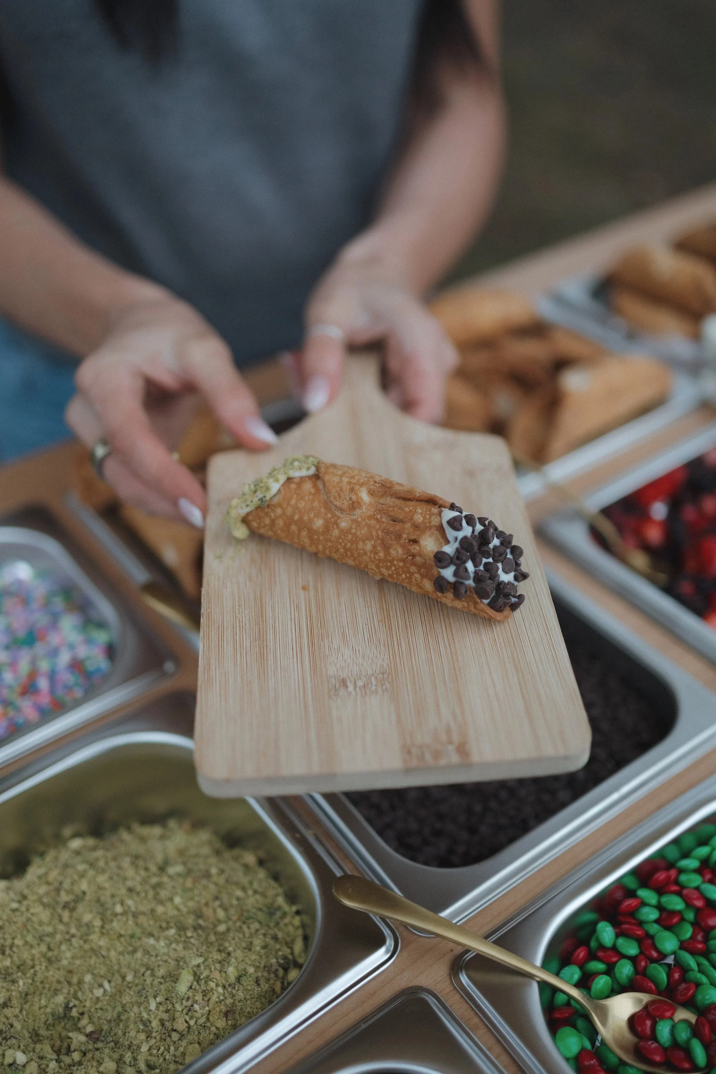 A person holding a wooden board with a fried pastry topped with green and white sprinkles and dark chocolate chips. In the background, there are containers with colorful sprinkles, crushed cookies, red and green candies, and other toppings for desserts.