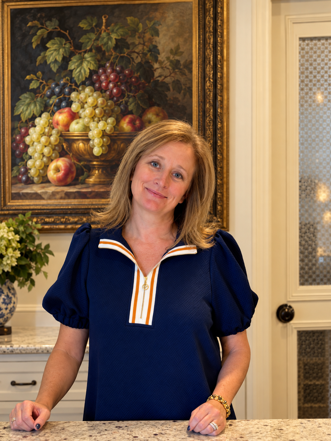 A woman with shoulder-length blonde hair, wearing a navy blue dress with puffed sleeves and a zipper detail, stands in a kitchen with a granite countertop. Behind her, there is a large framed painting of a bowl of assorted grapes and apples. To the left, a potted plant with white flowers is visible, and to the right, a door with decorative frosted glass panels.