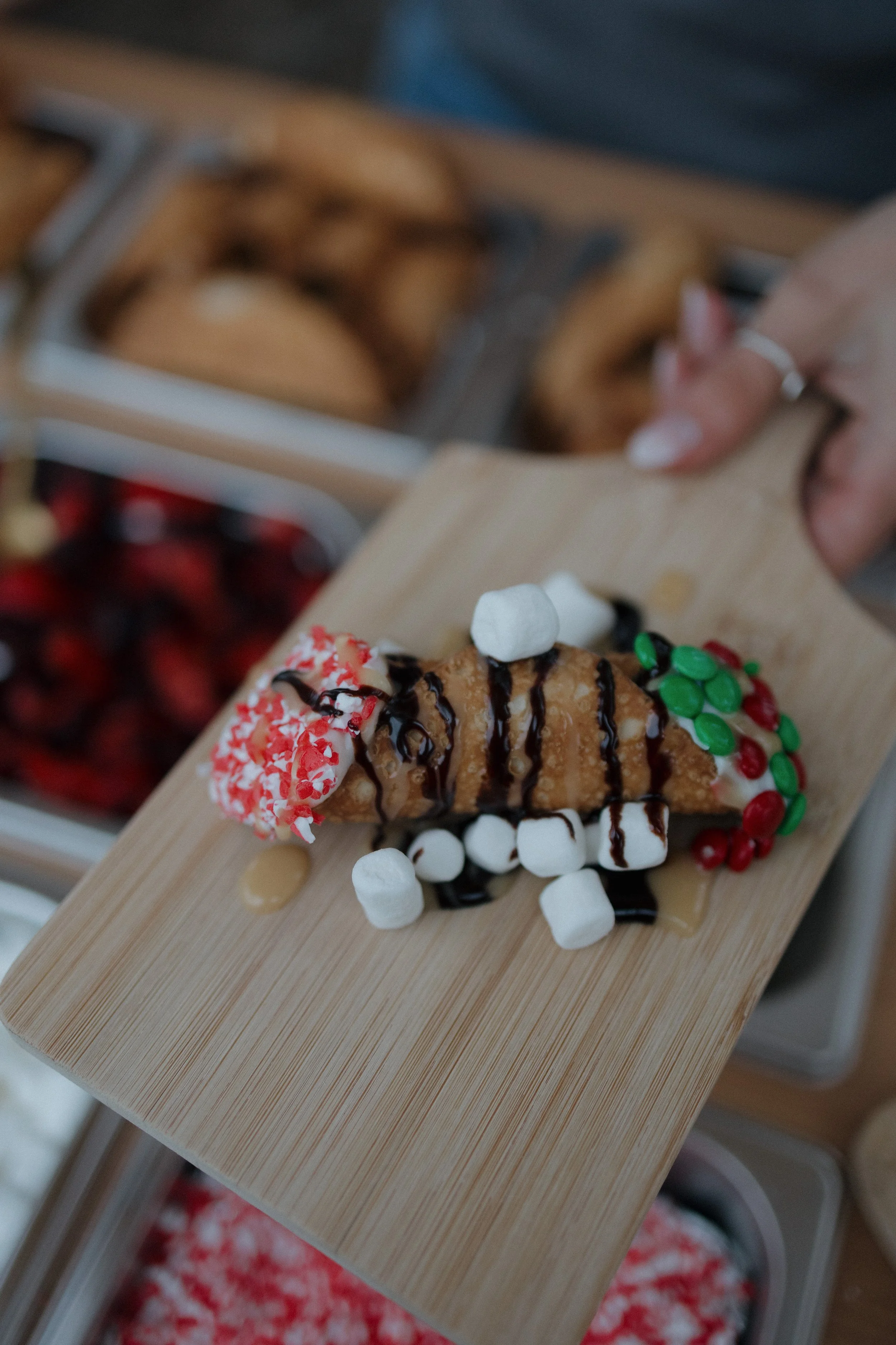 A slice of Christmas-themed dessert cake with white icing, red sprinkles, green and red candies, chocolate drizzle, and marshmallows on a wooden tray.